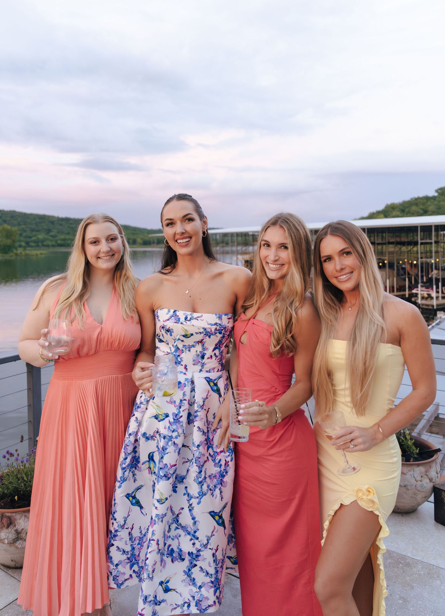A group of friends in summer dresses pose together at a lakeside dock during sunset.