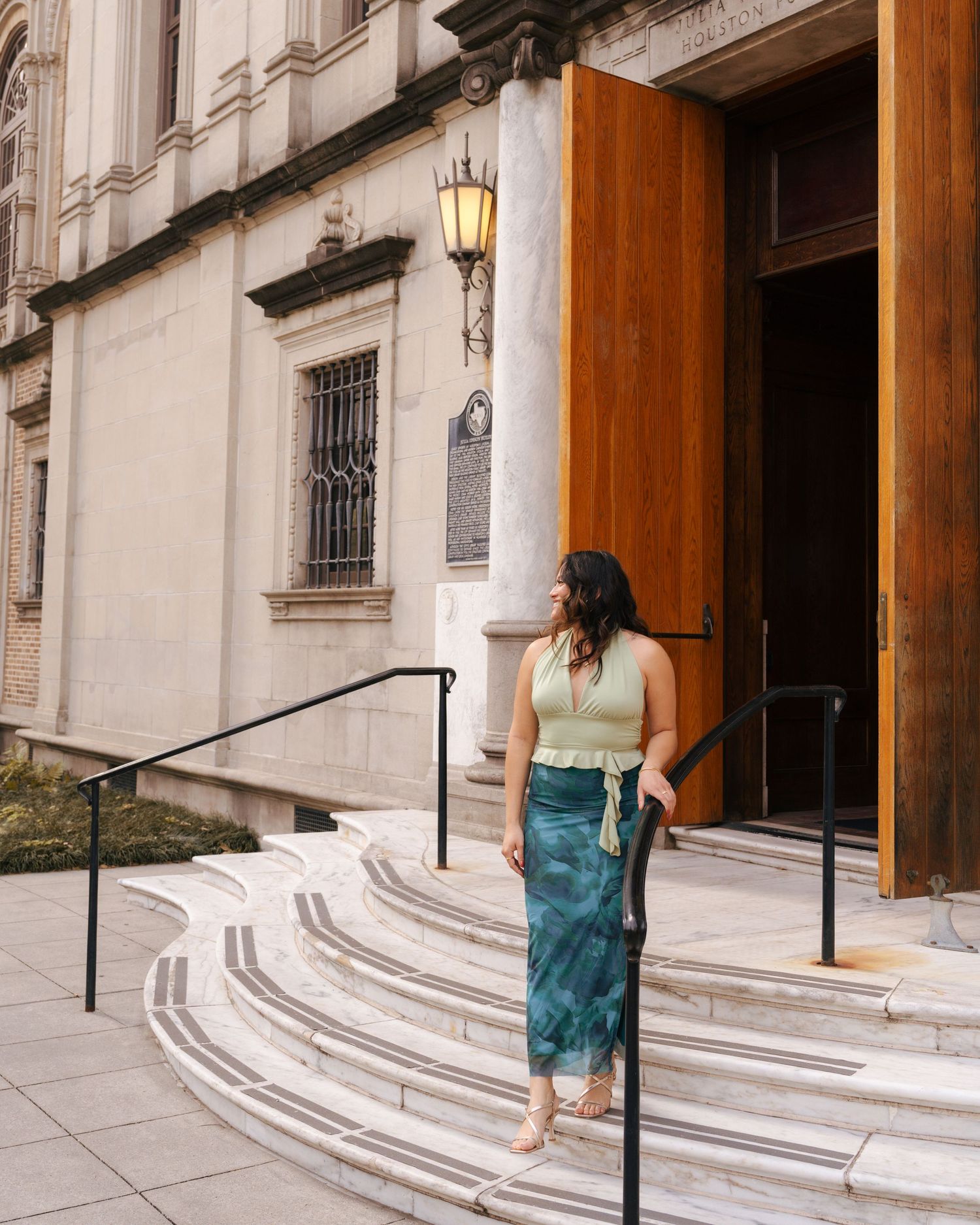 Woman in sleeveless top and teal skirt stands on curved stone steps of historic building with vintage lamp and wooden door.