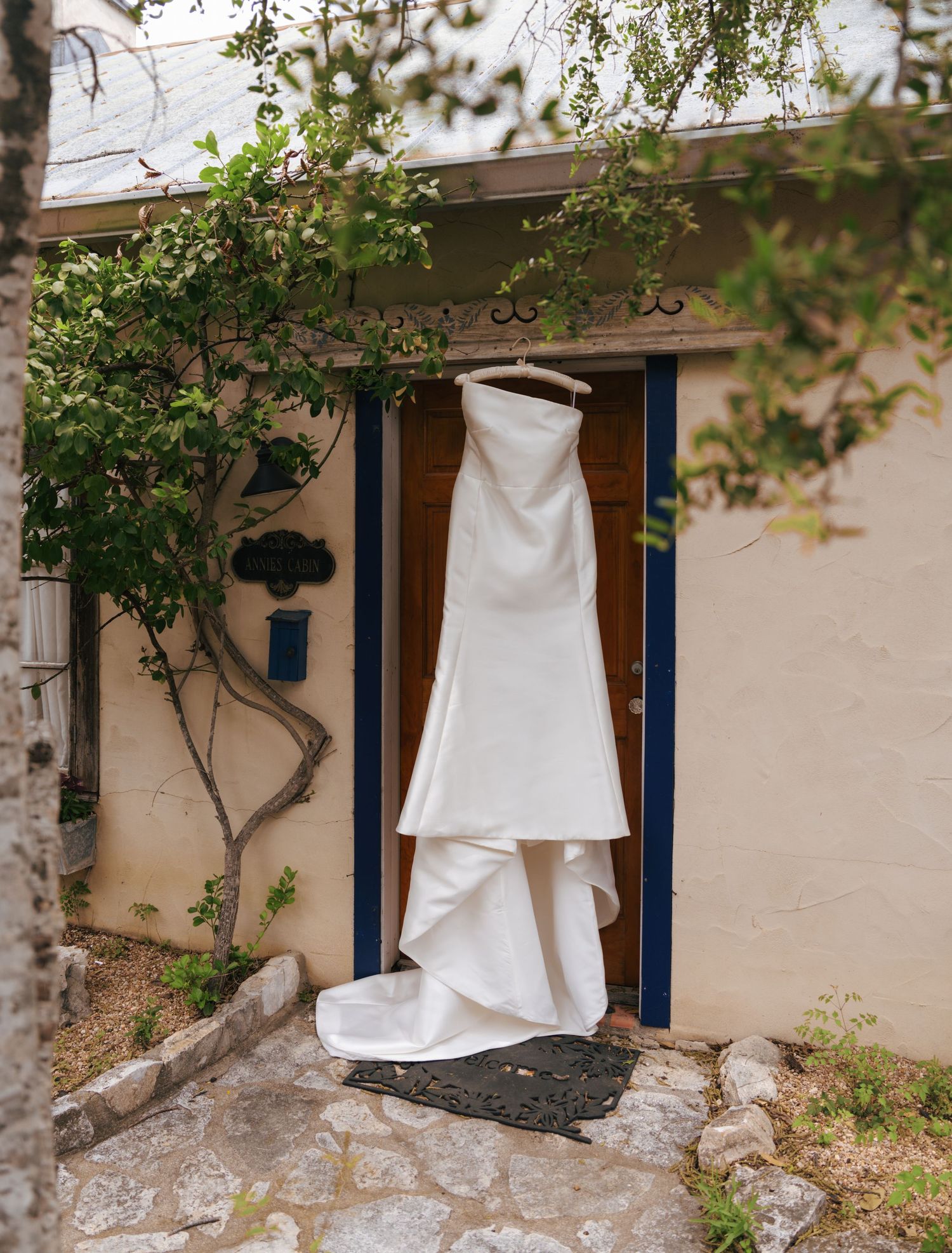A white wedding dress hangs elegantly against a rustic wall with climbing vines and blue accents.