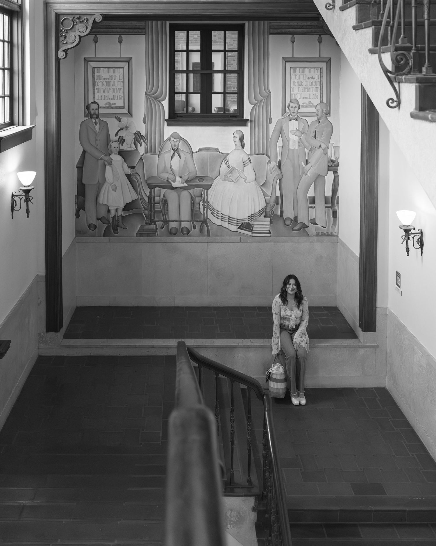 A black and white photograph shows a stairway landing with a decorative wall mural depicting a colonial-era social scene.