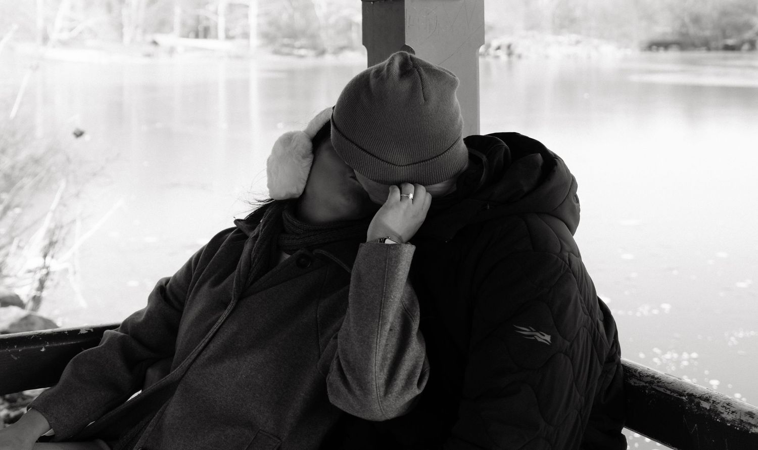 Two figures embrace near a lake with tall buildings visible in the background.