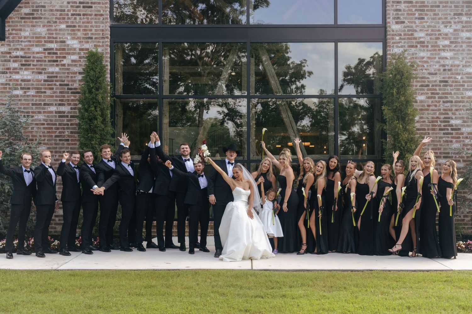 Large wedding party in formal attire poses together against a brick wall backdrop.