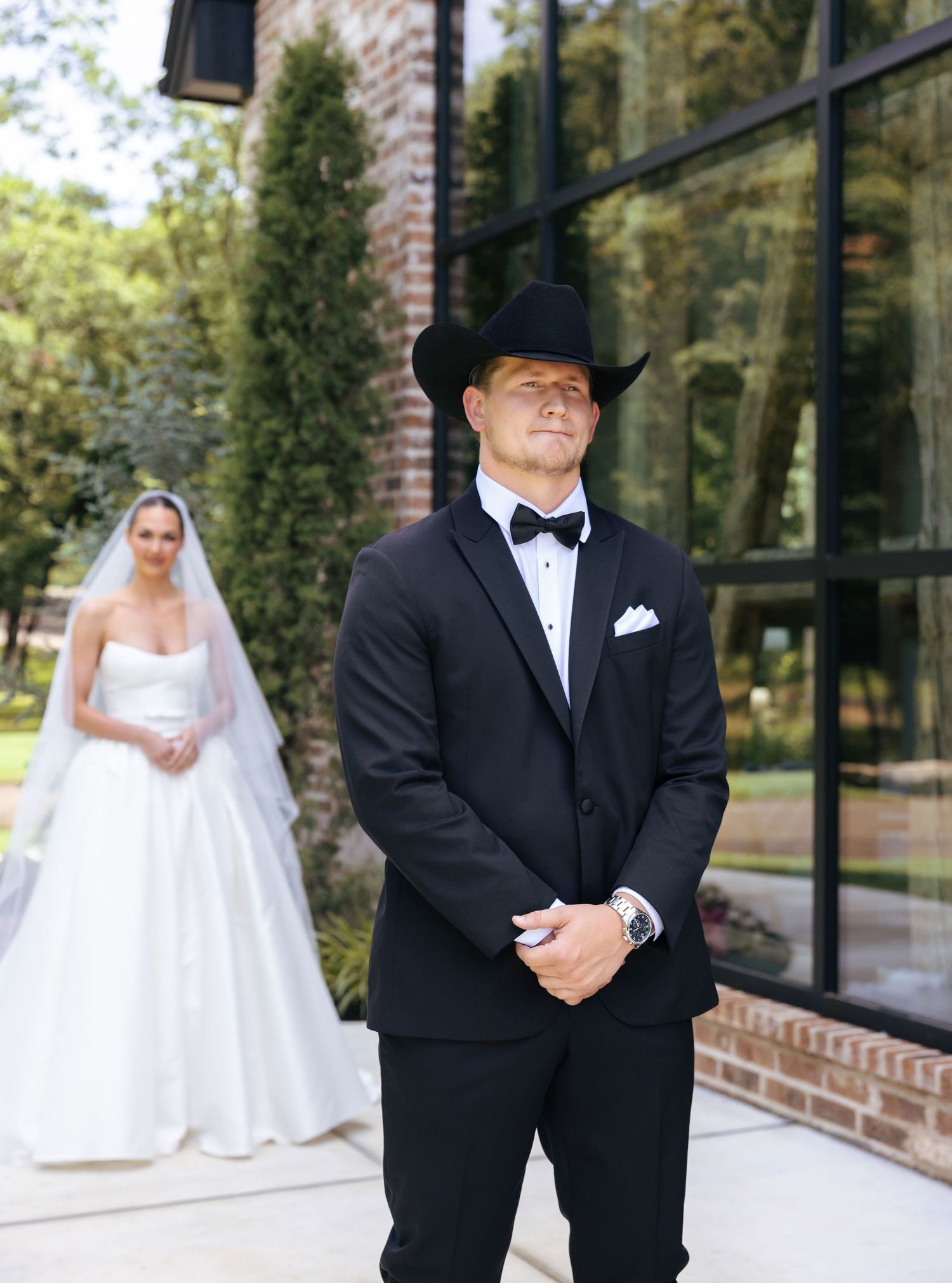 Wedding scene with groom in black tuxedo and cowboy hat standing in front of glass building.
