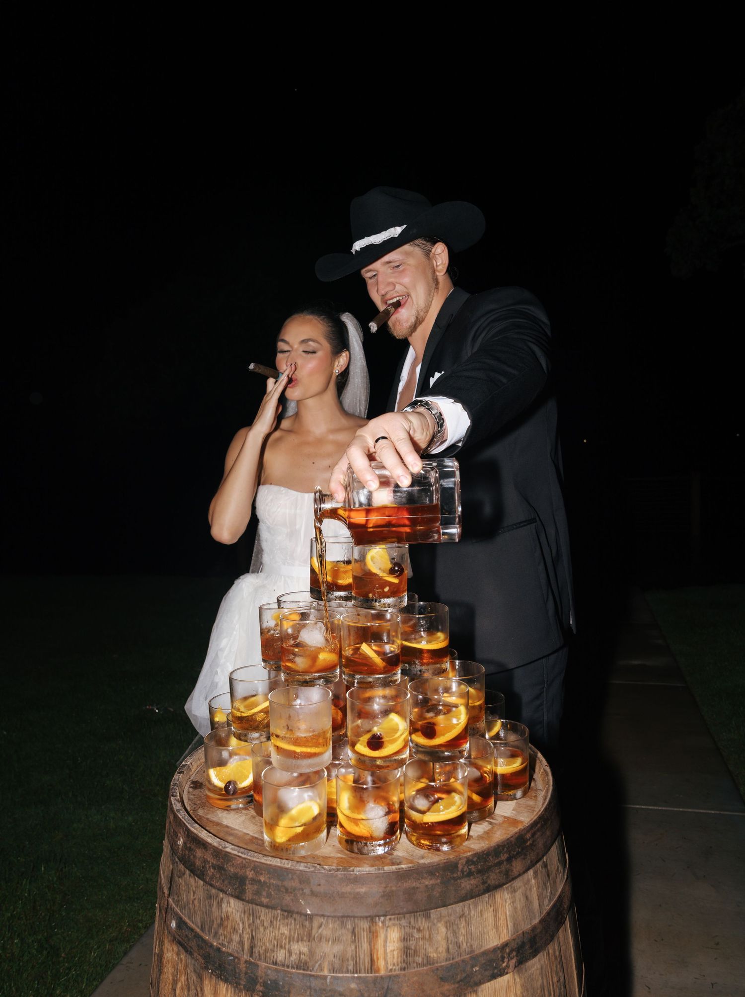 Wedding couple near a wooden barrel display of glowing cocktails.