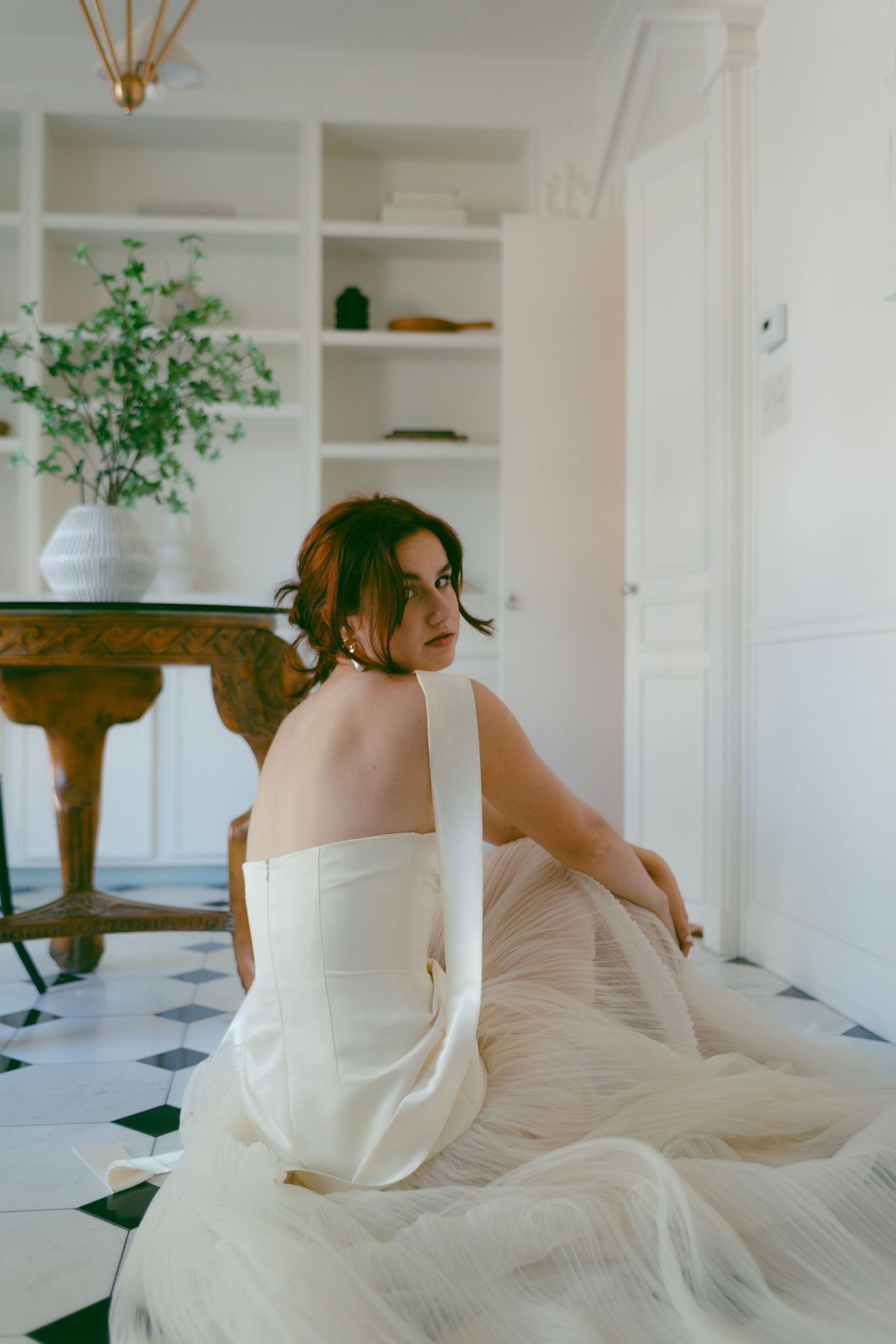 A white dress billows dramatically against white built-in shelving and green plants.