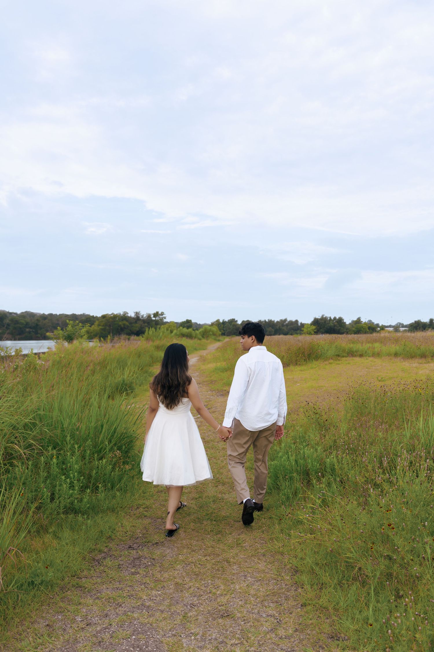 Two people walking together on a dirt path through a grassy meadow on a cloudy summer day.