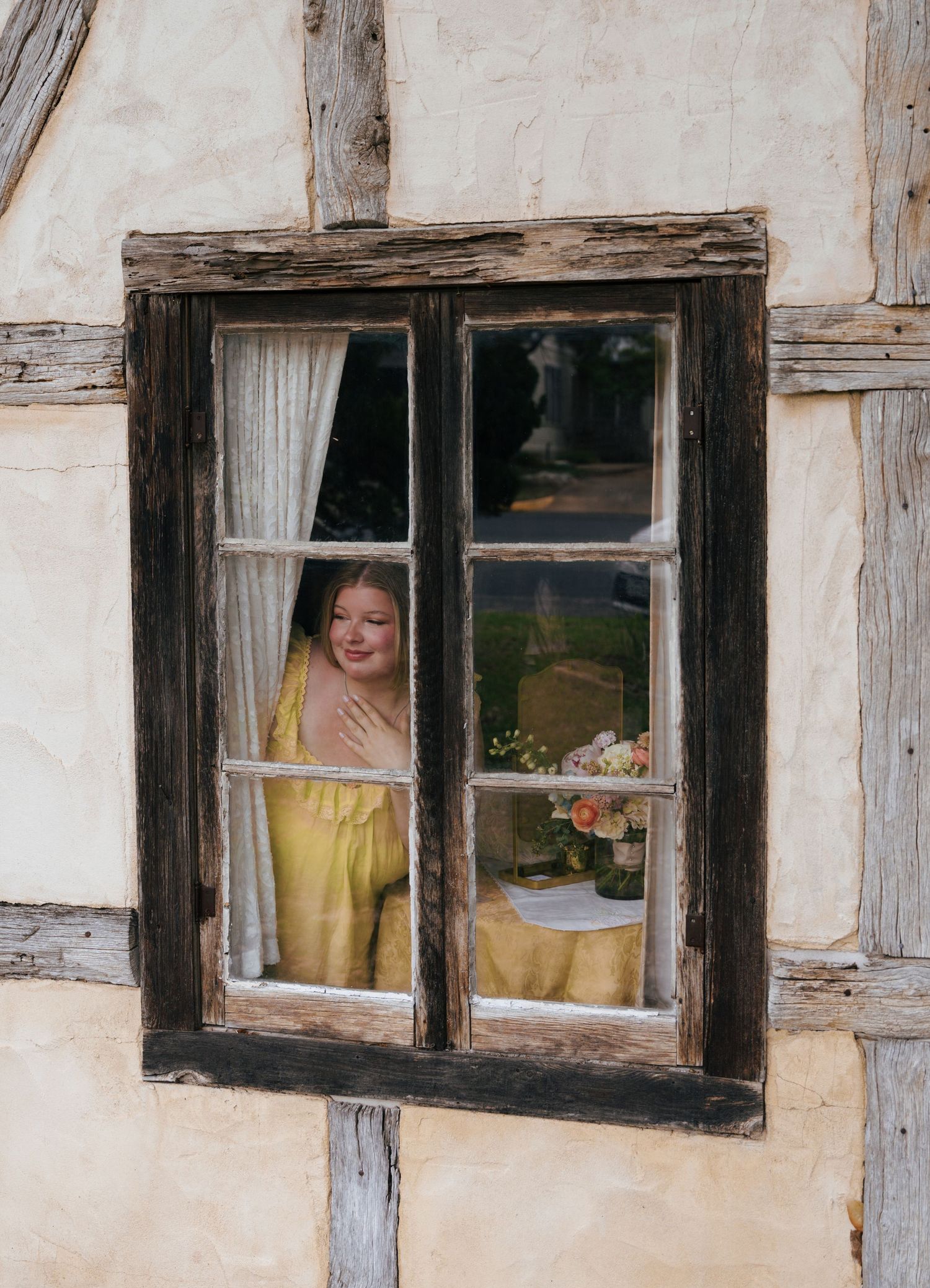 A rustic window with wooden frame and exposed timber beams shows a yellow interior with flowers on a table.