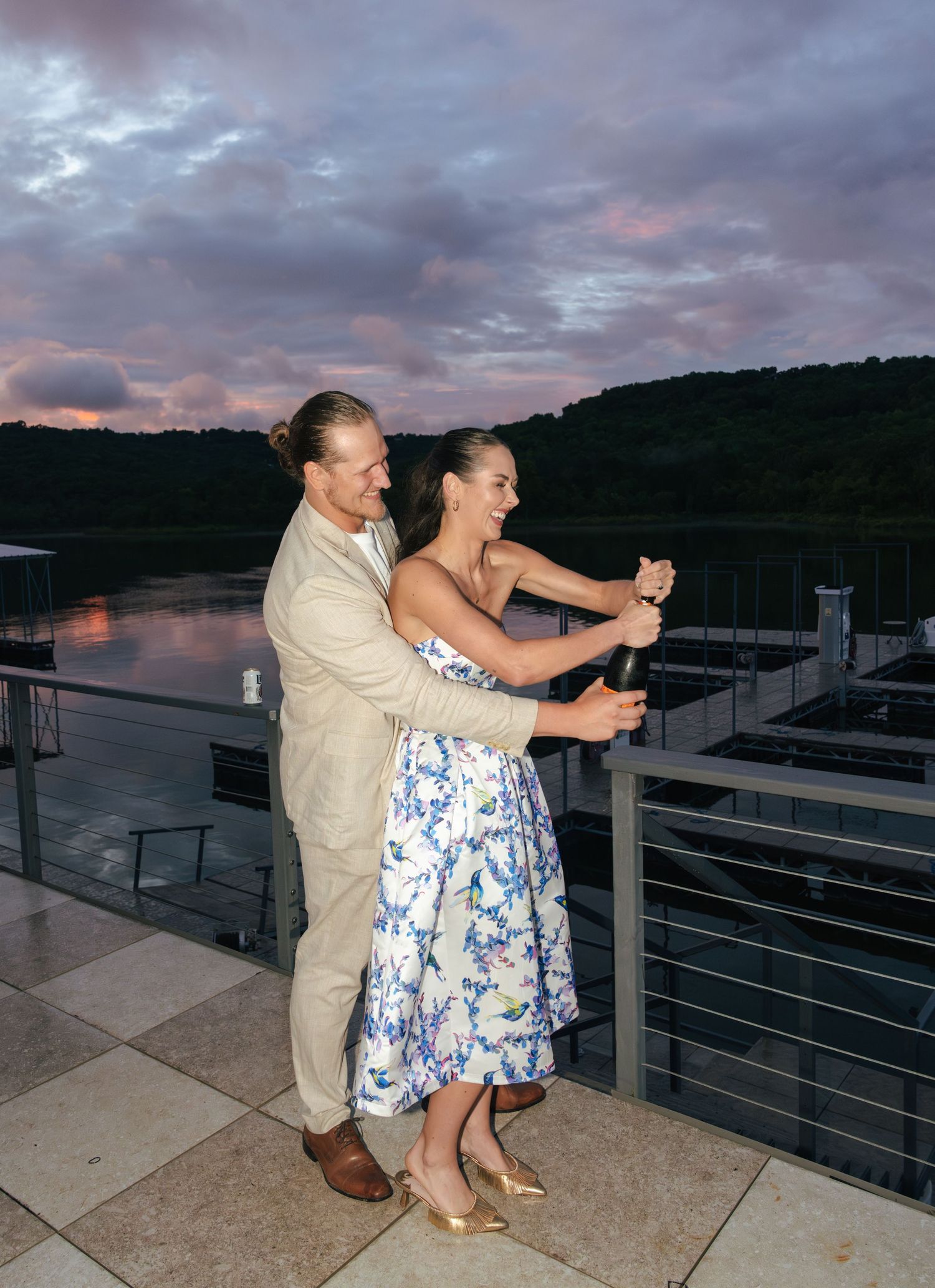 A couple shares a romantic moment dancing together on a marina dock at sunset with boats in the background.