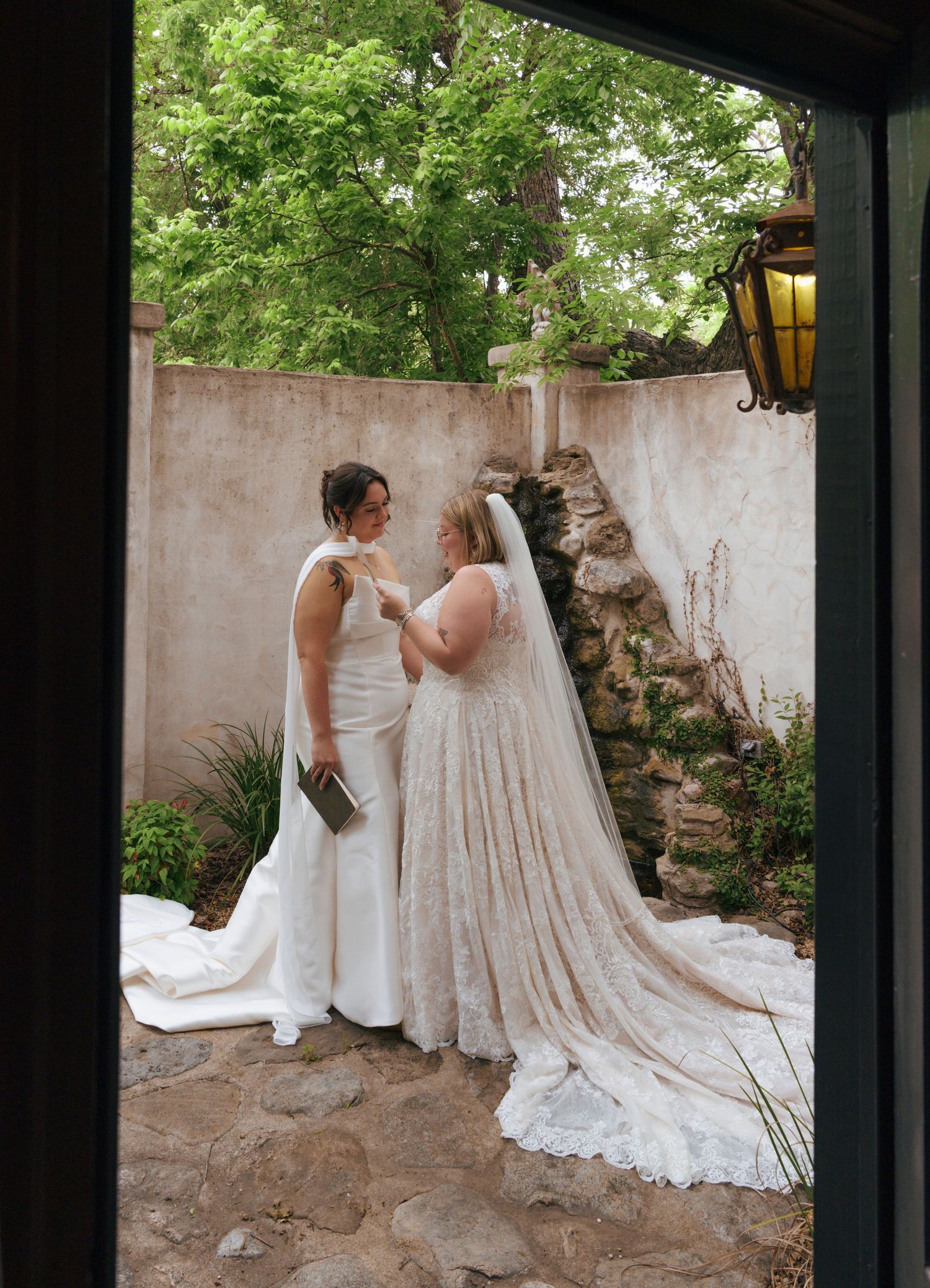 Two people in elegant white dresses share intimate moments in a garden courtyard with stone walls and lanterns.