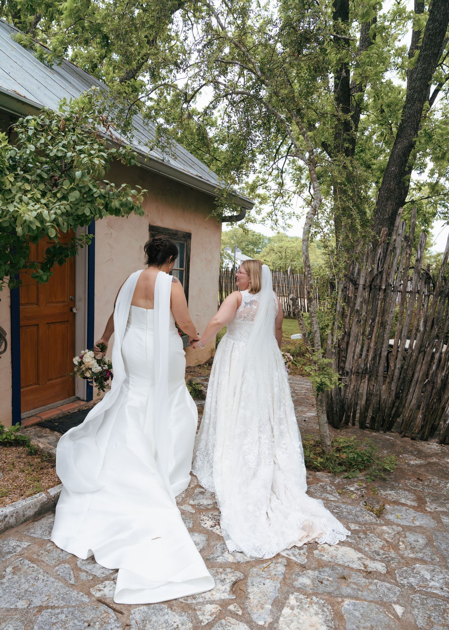 Two figures in elegant white wedding dresses walking together on a rustic outdoor path surrounded by trees and wooden fencing.