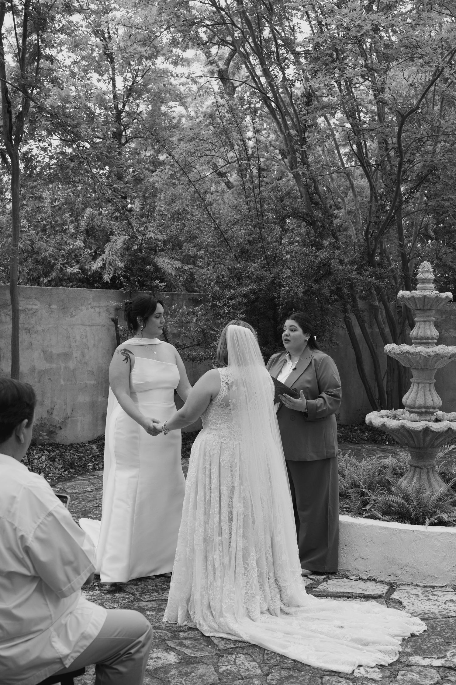 Black and white wedding scene in a garden with elegant white dresses and formal attire.