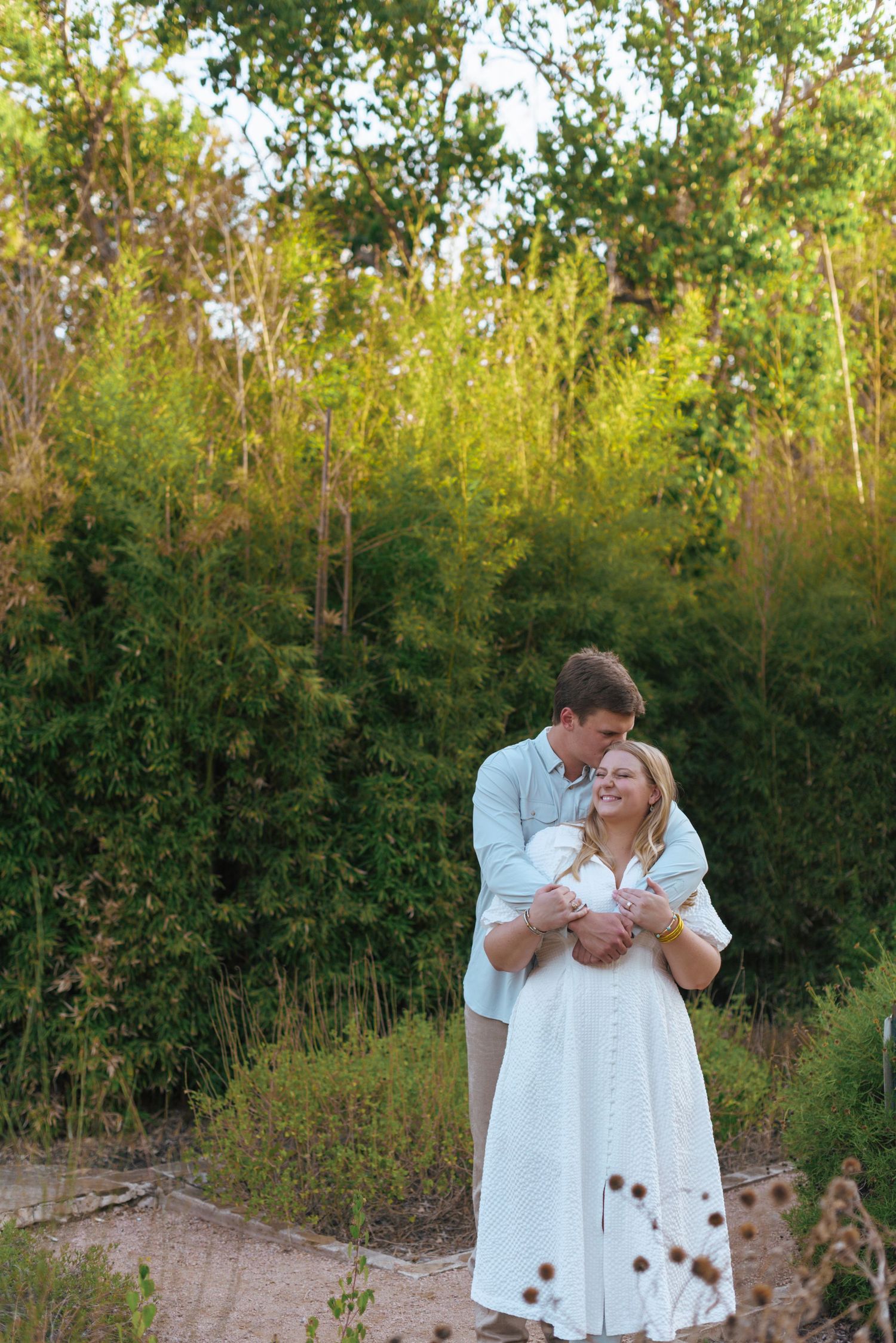 A couple in white outfits embraces tenderly in a natural outdoor setting with green foliage in the background.