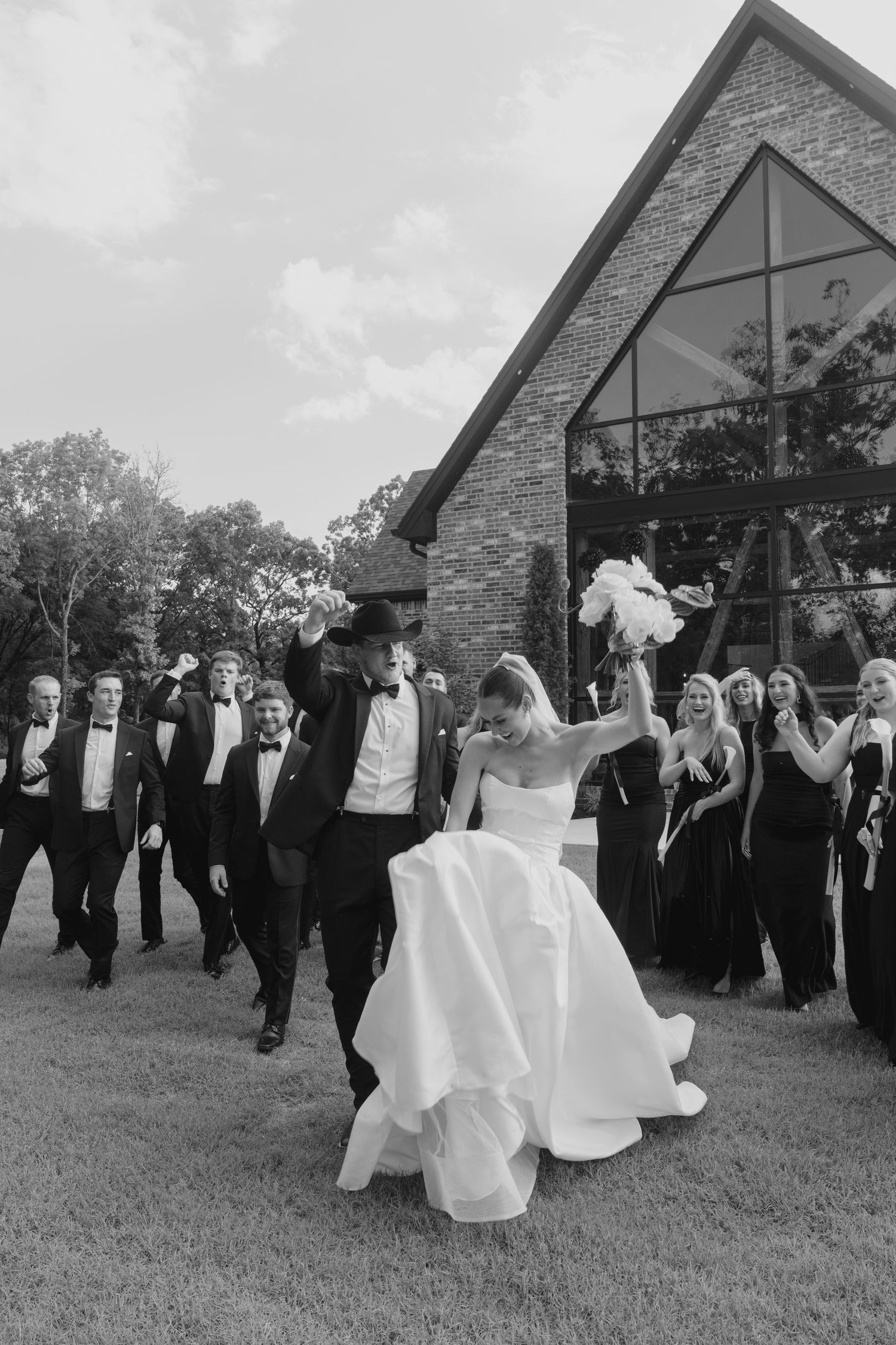 Wedding party in formal attire performs choreographed dance moves on grass near stone building.