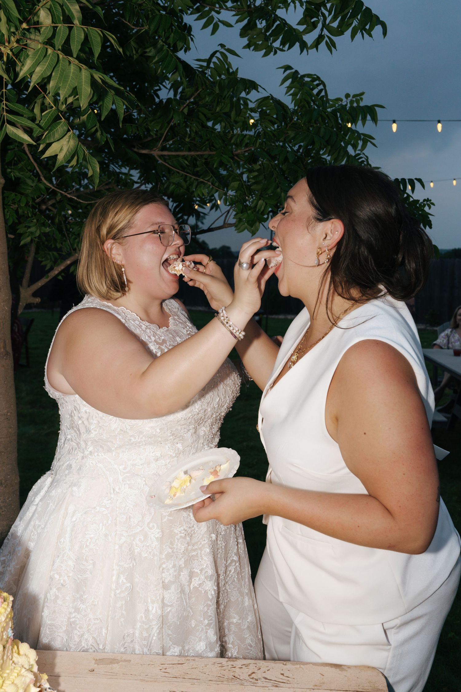 Wedding couple playfully feed each other cake during their reception celebration outdoors under string lights.
