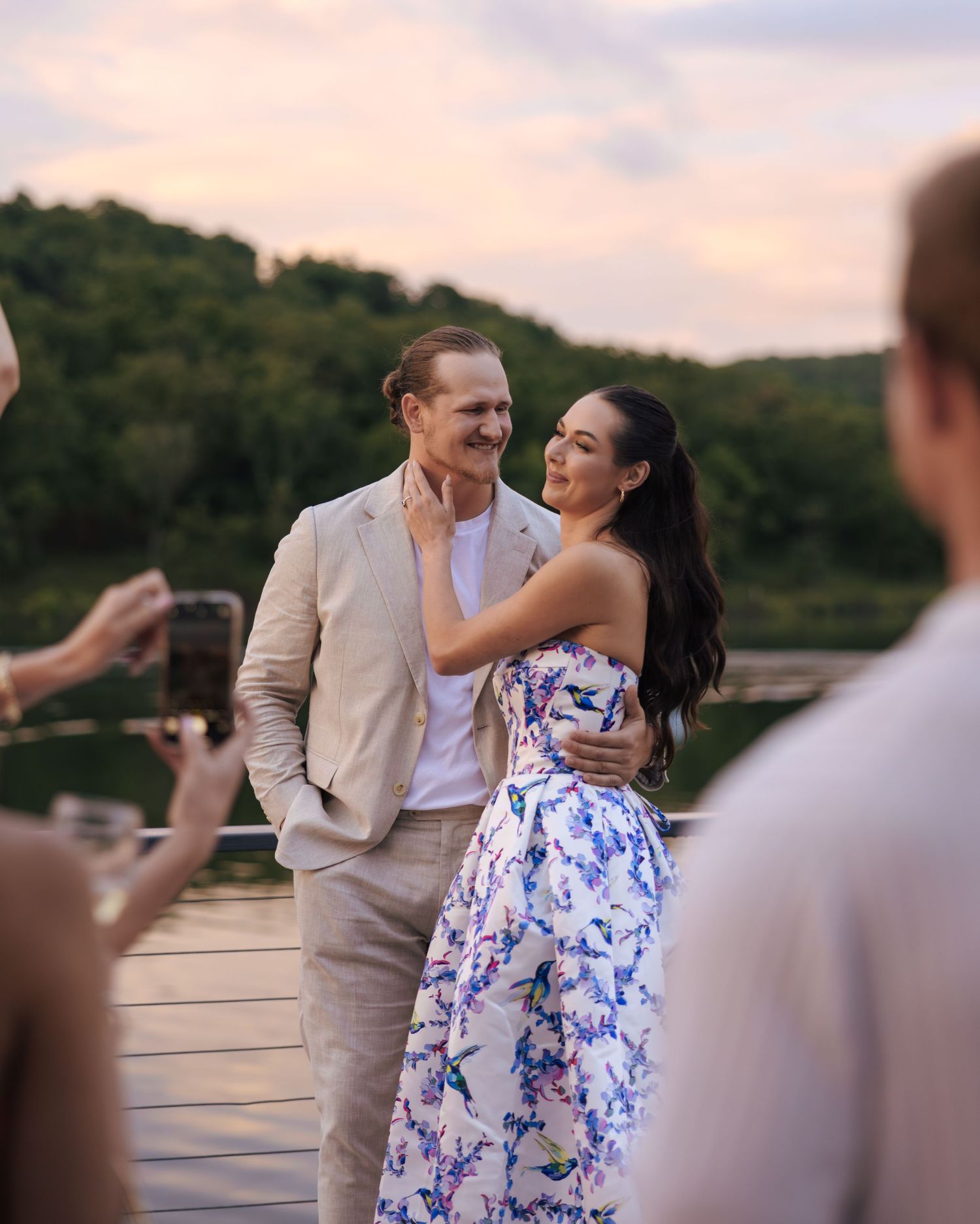 A couple in formal attire share a romantic dance on a wooden deck at sunset with blurred onlookers in the background.