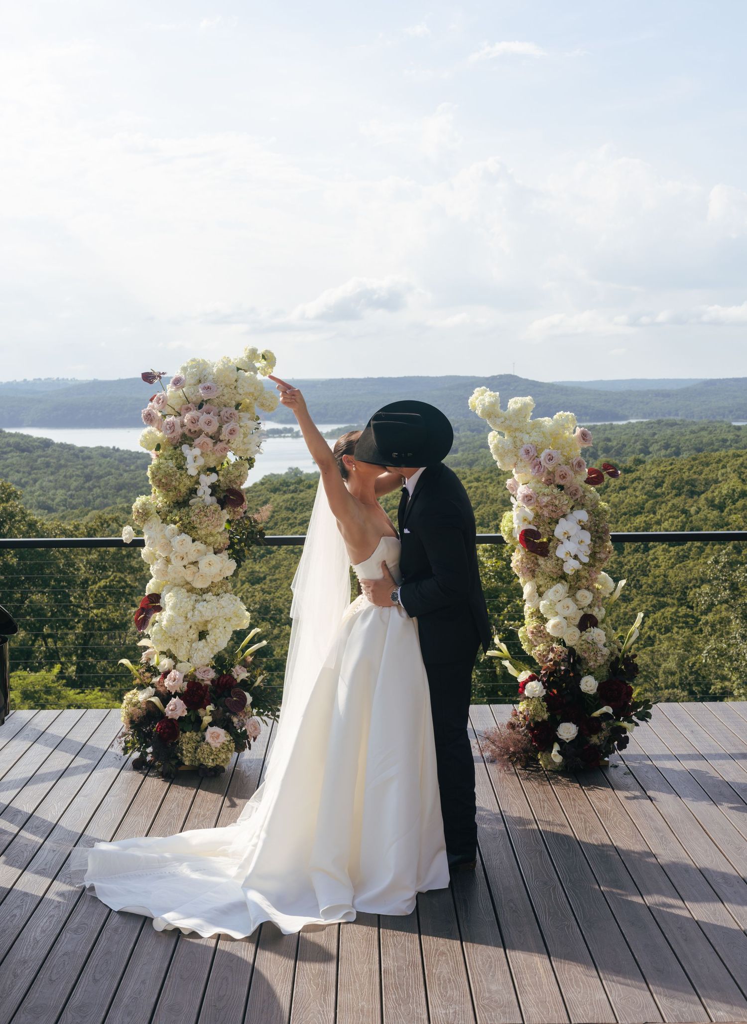 A wedding couple poses on a wooden deck overlooking mountains with elegant white floral arrangements.