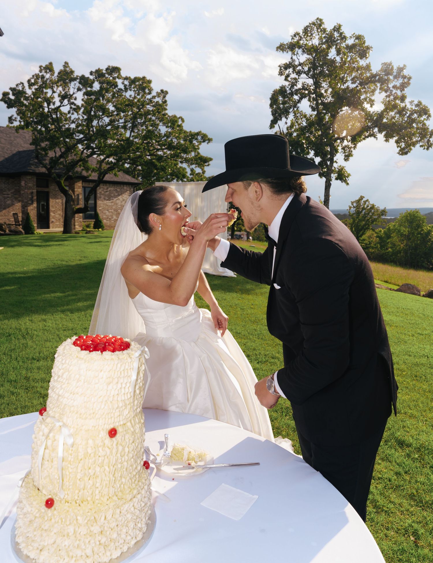 A playful moment between newlyweds sharing cake beside their elegant white wedding cake with red accents.