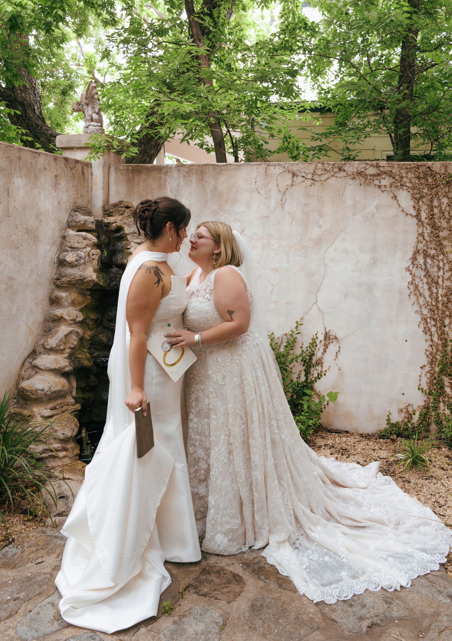 Two brides in white dresses share intimate moments against a garden wall with ivy.