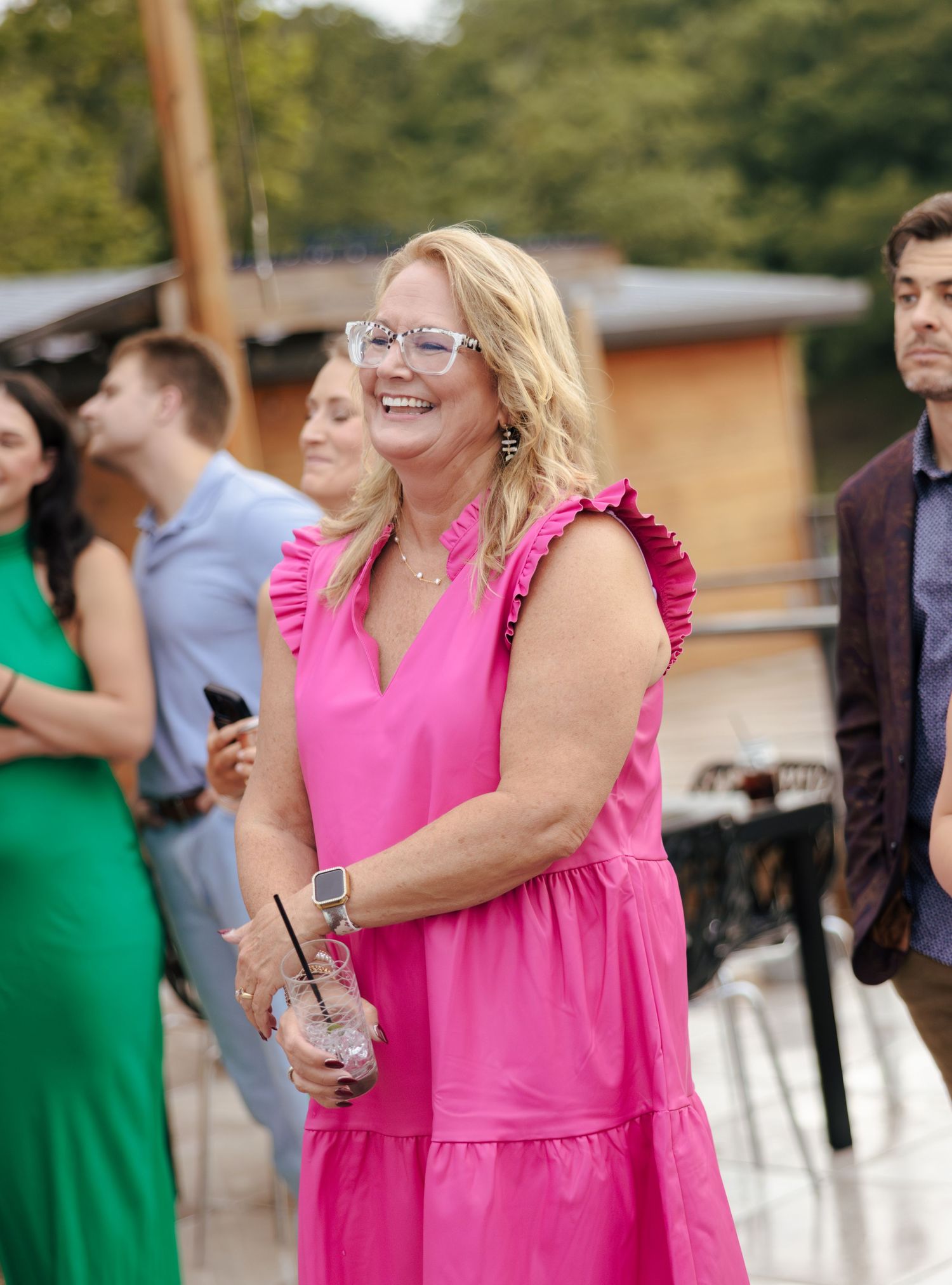 Woman in bright pink dress laughs joyfully during outdoor celebration with guests in background.