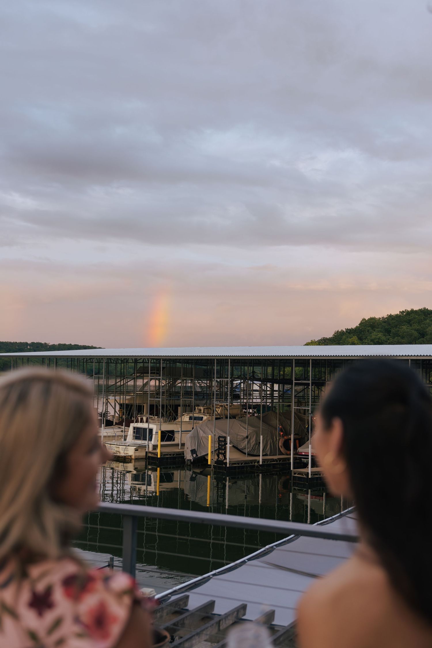 People gathered on a rooftop deck at sunset with a marina and rainbow visible in the cloudy sky.
