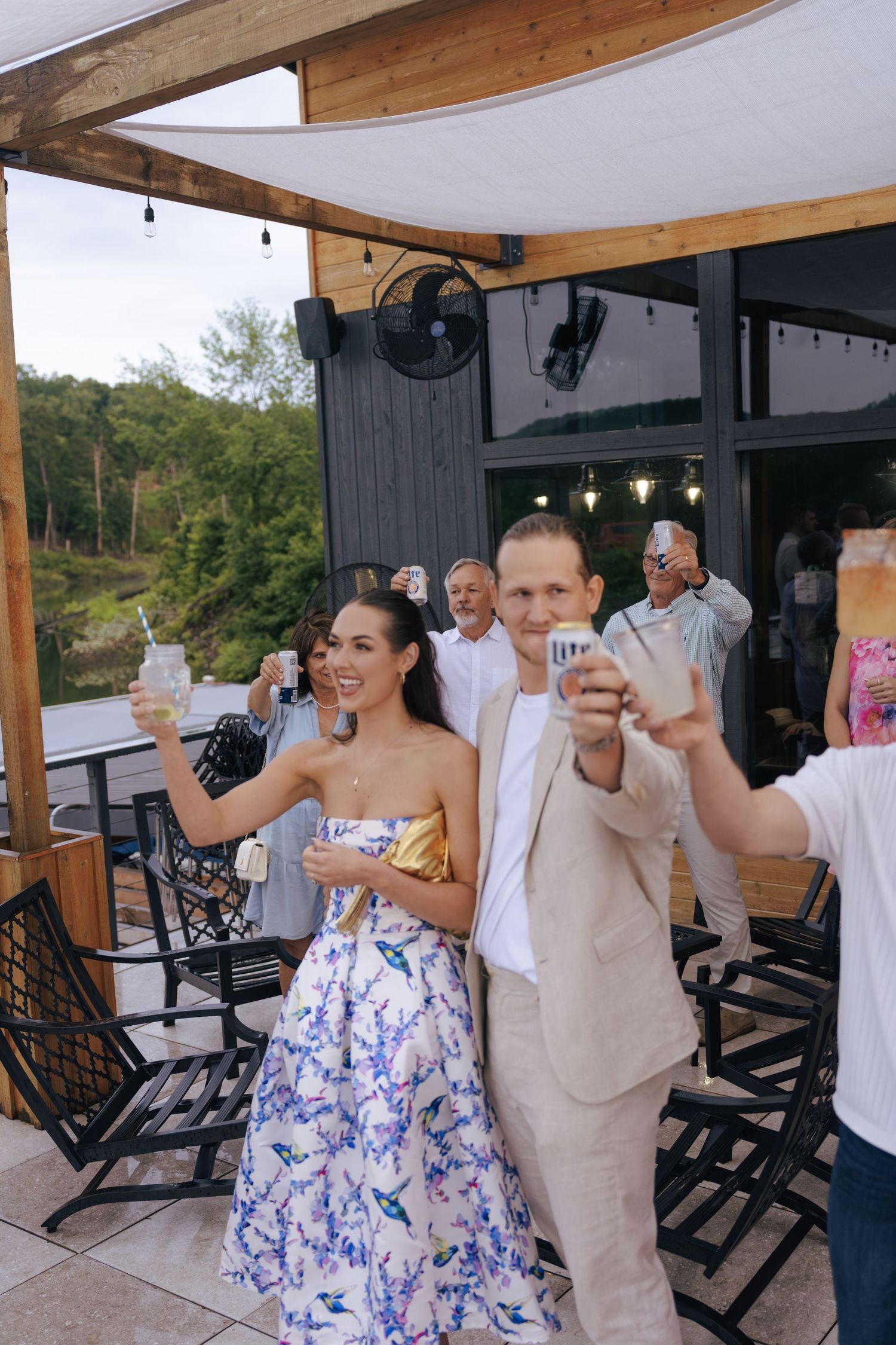 Wedding guests raise their glasses in a toast on an outdoor covered patio during a celebration.