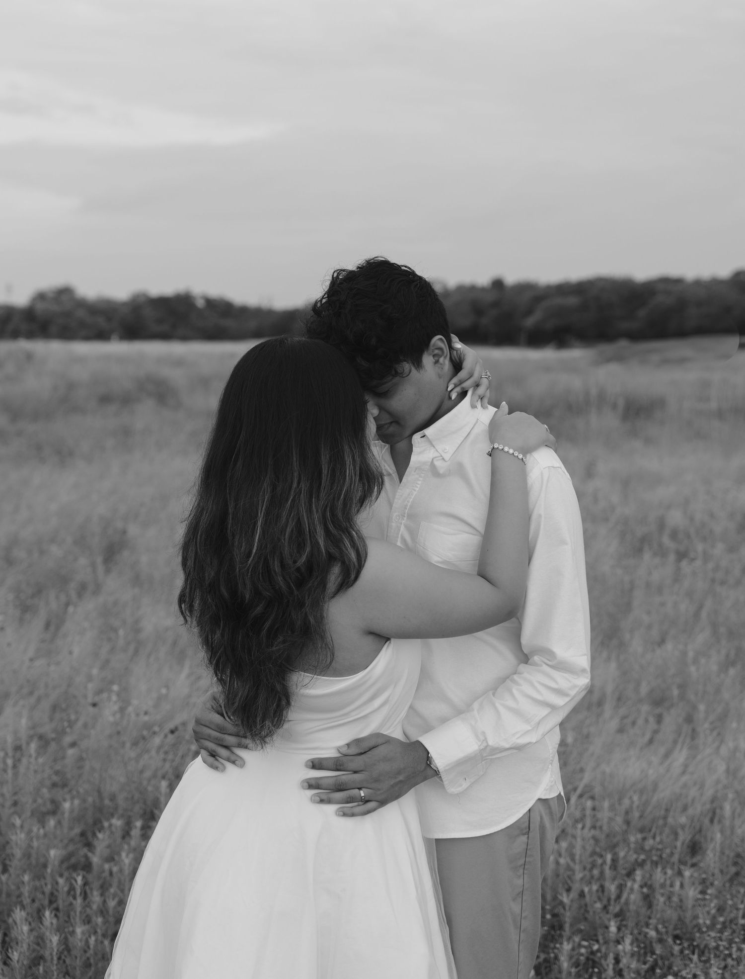 A romantic couple embraces in a serene meadow field with tall grass at sunset, captured in black and white.