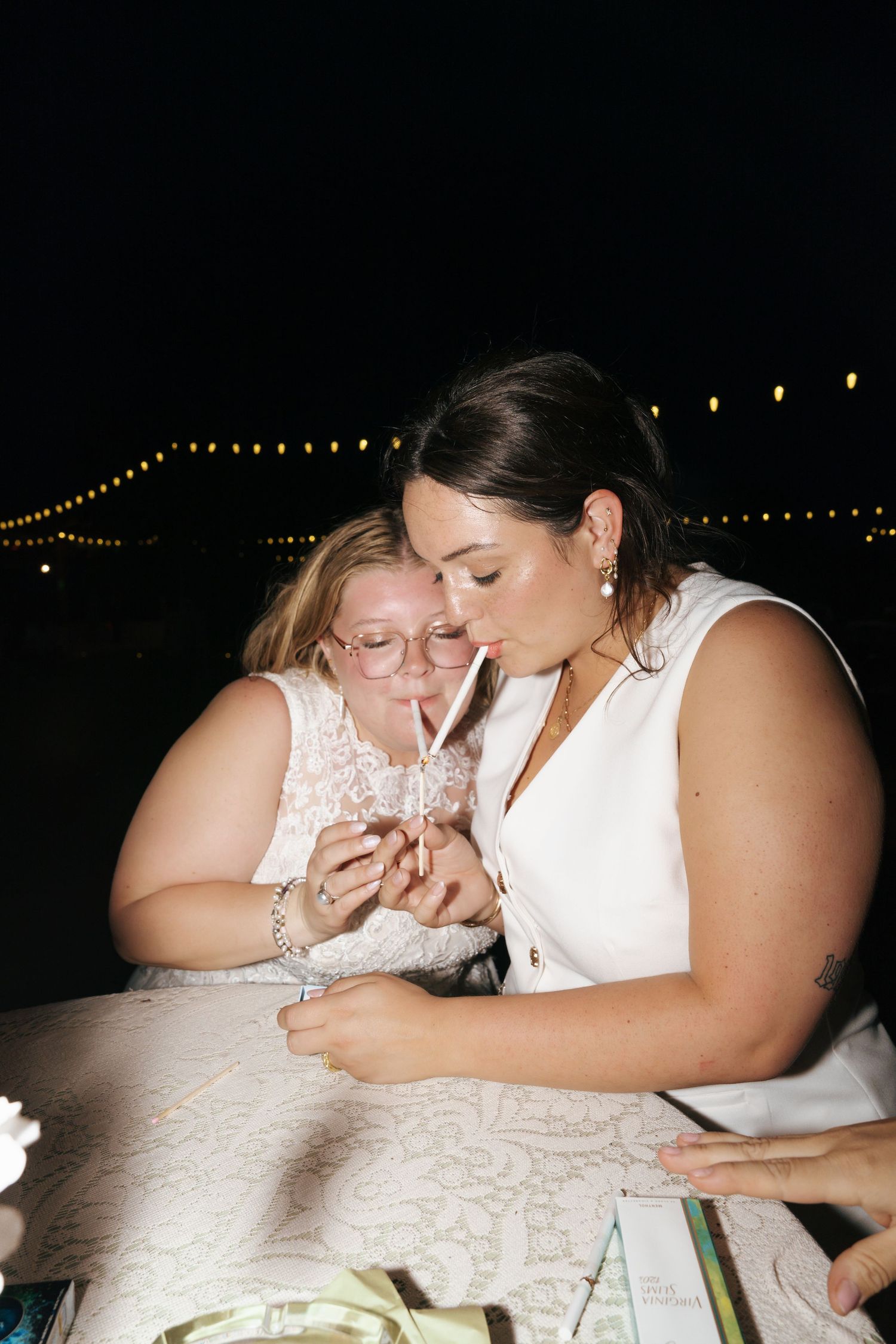 Two people in white dresses share an intimate moment while looking at something together at a candlelit table.