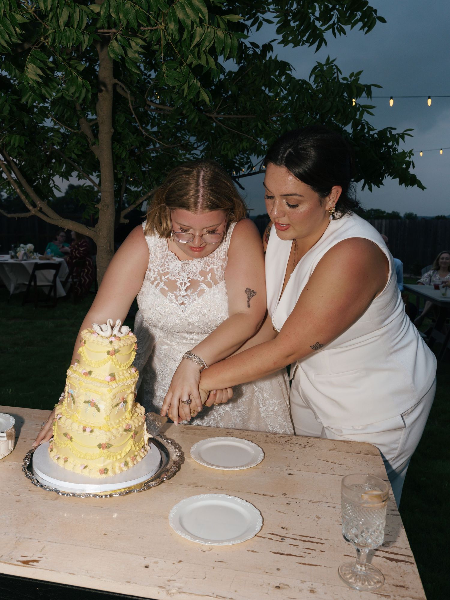 Two people in wedding attire cut into a three-tiered yellow and white wedding cake on a rustic wooden table outdoors.