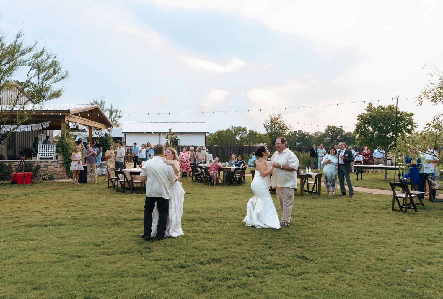 Wedding guests gather on a grassy lawn for an outdoor celebration with white chairs and tents at sunset.