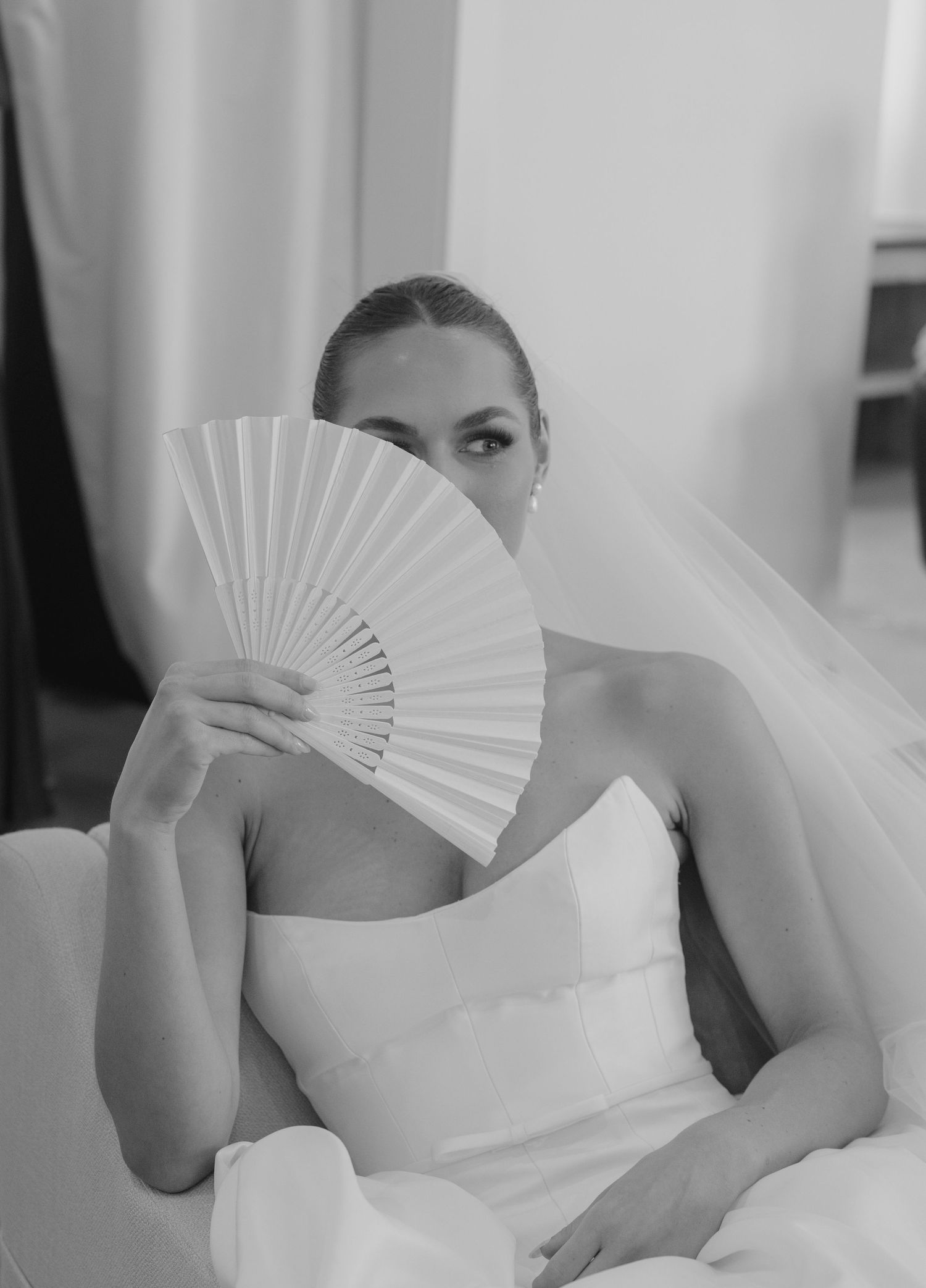 Bride in white dress poses with decorative fan in minimalist setting.