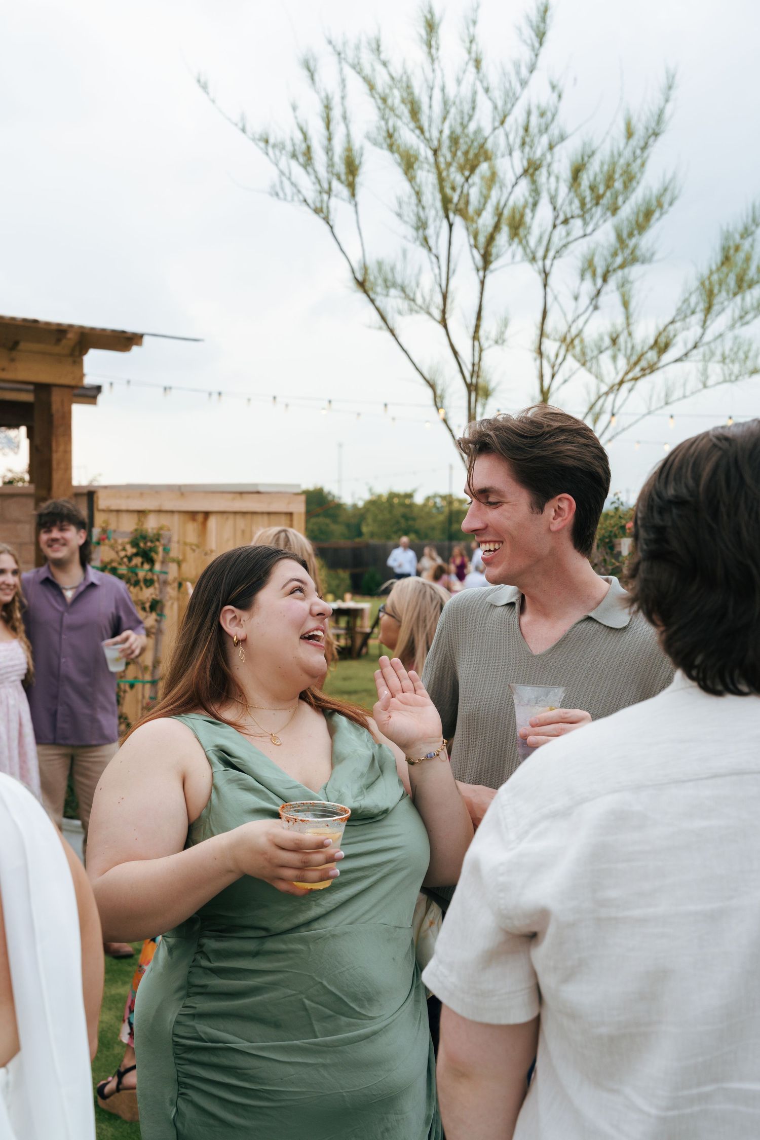 People enjoying drinks and conversation at an outdoor party with string lights and trees in the background.