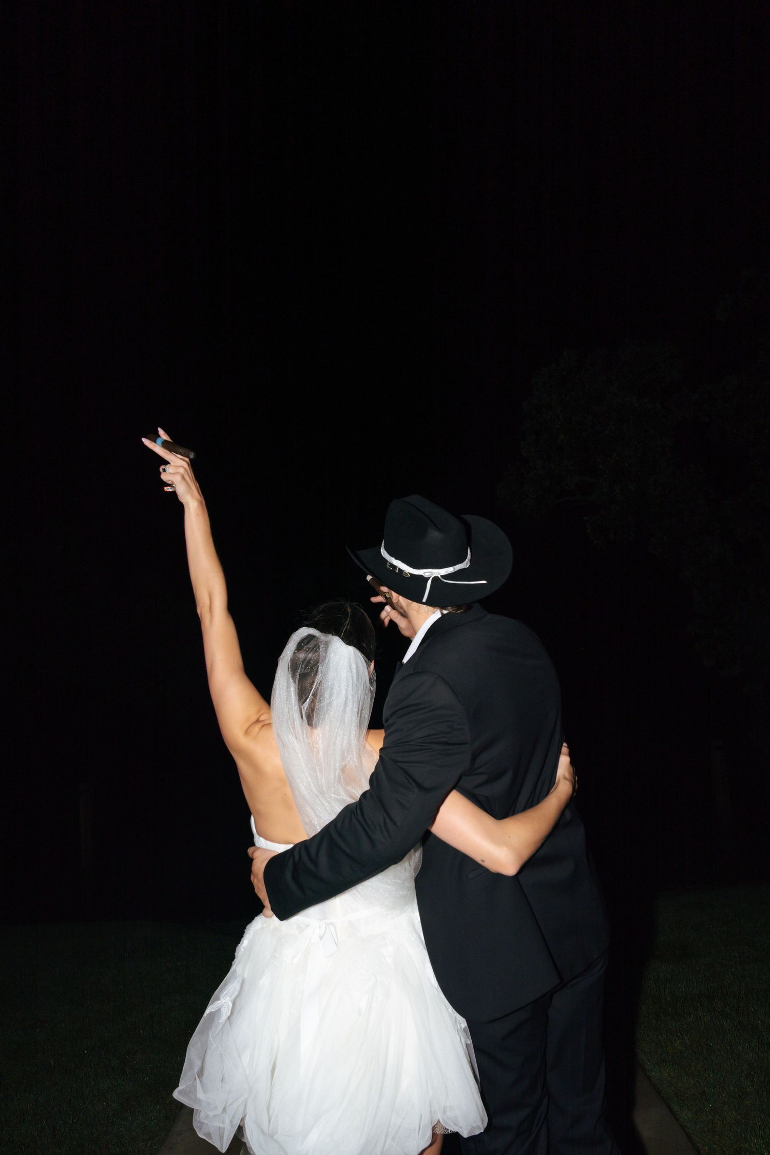 A couple in wedding attire smoking together against a dark background.