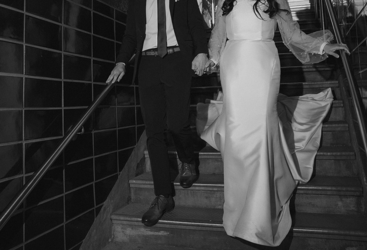 A couple descending elegant stairs in formal attire with a white gown and dark suit visible in black and white.