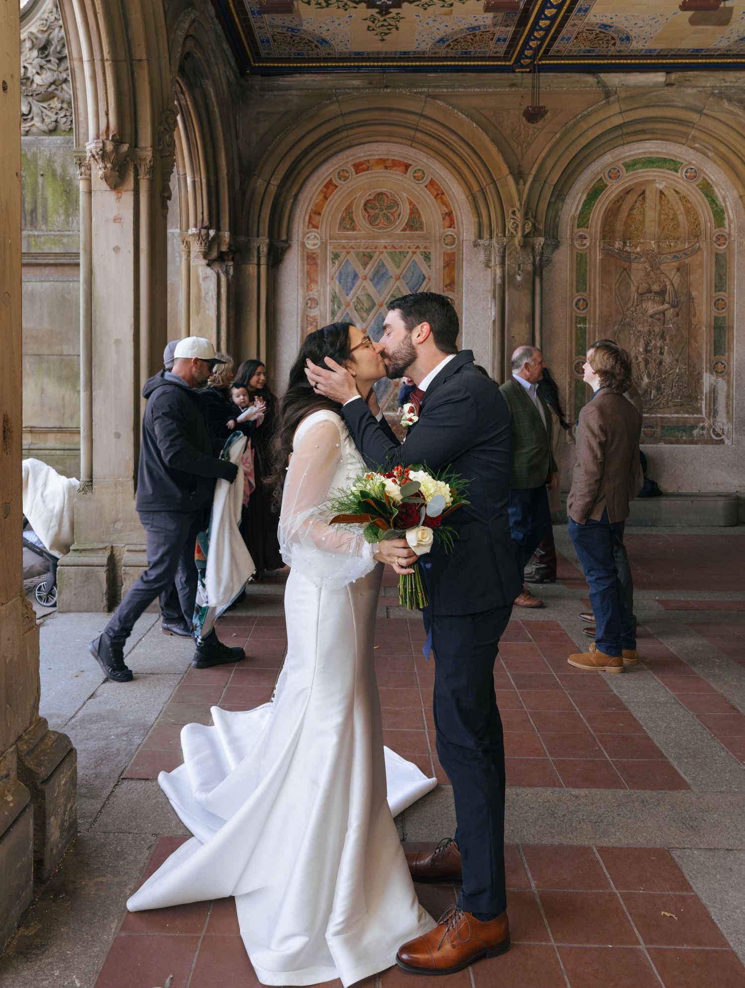 A wedding ceremony takes place in an ornate historic colonnade with arched ceilings and decorative details.