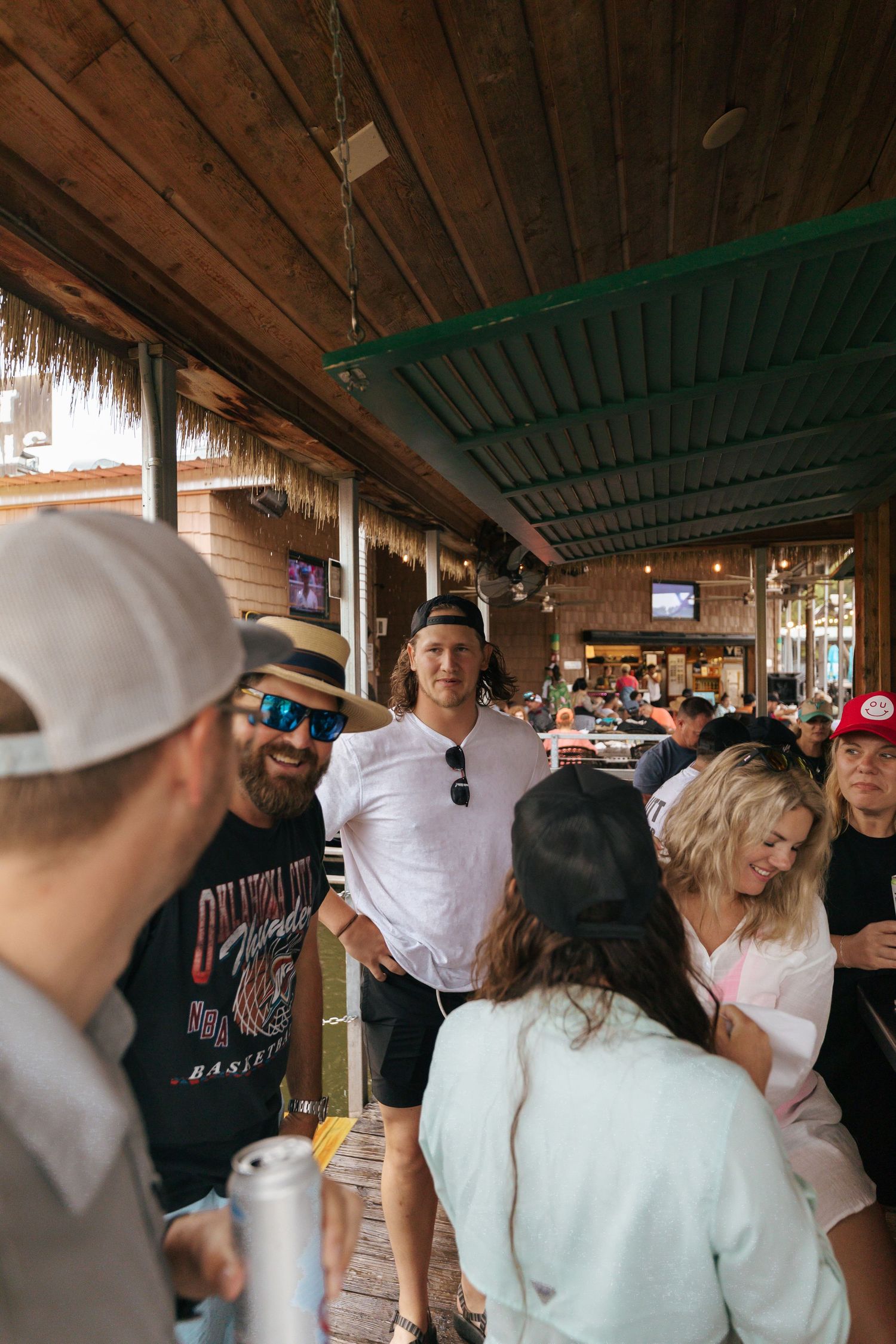 Group of people socializing and drinking at a casual tiki bar with wooden beams and thatched roof decor.