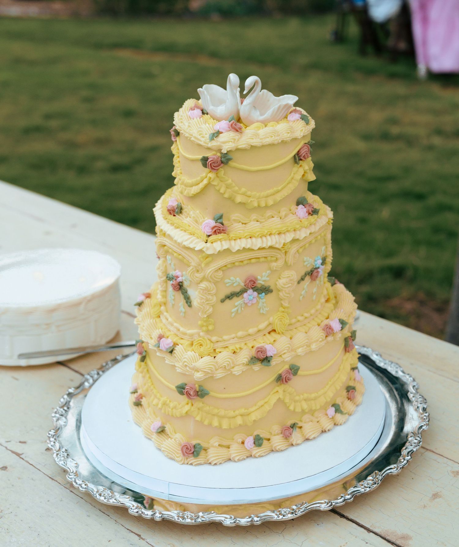 A three-tiered yellow buttercream wedding cake decorated with delicate piping and small purple flowers on a silver platter.