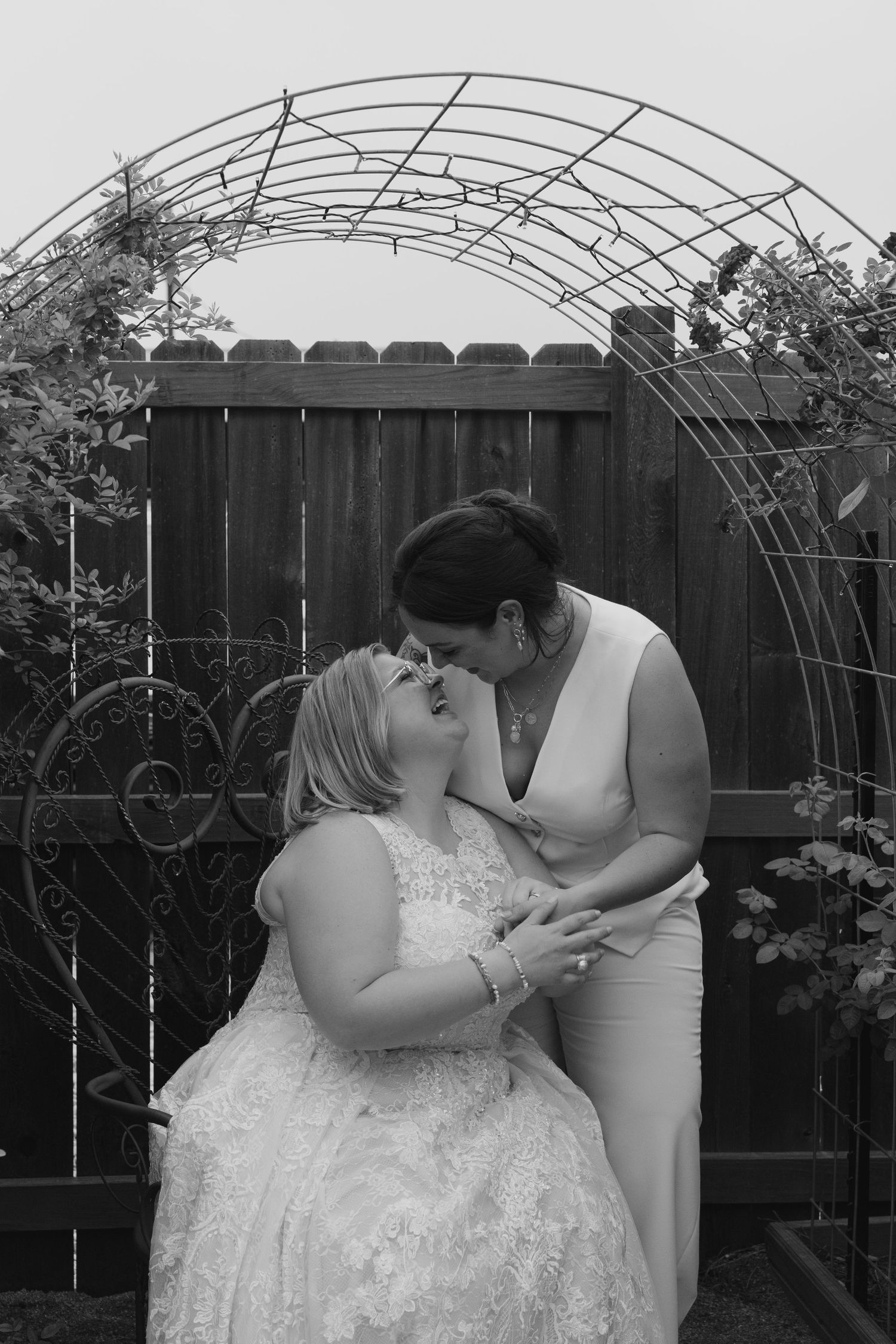 Two people in formal attire share an intimate moment in front of a garden trellis backdrop.