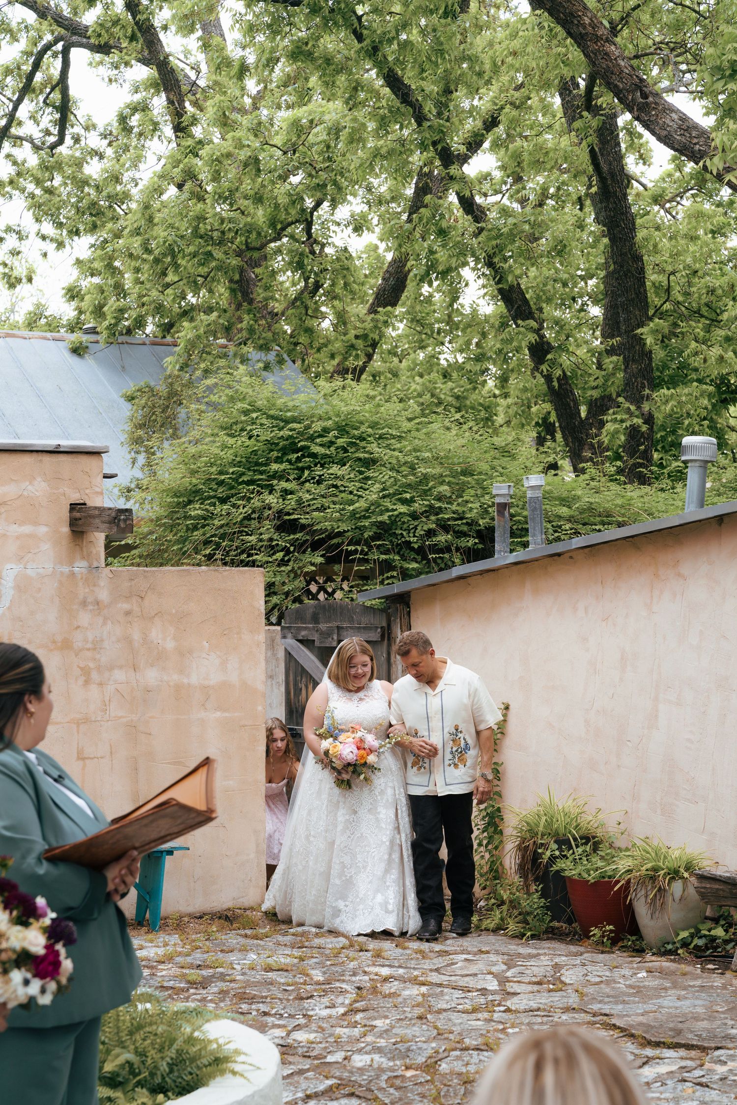 A wedding ceremony takes place in a courtyard garden with stone walls and lush green trees overhead.