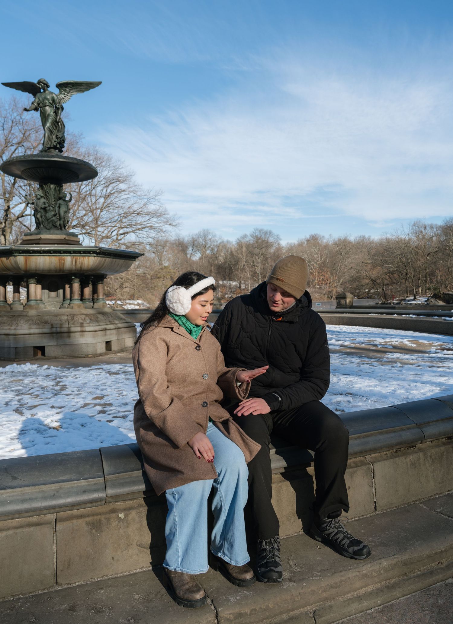 Two people sit together on a stone bench overlooking a frozen lake in Central Park on a clear winter day.