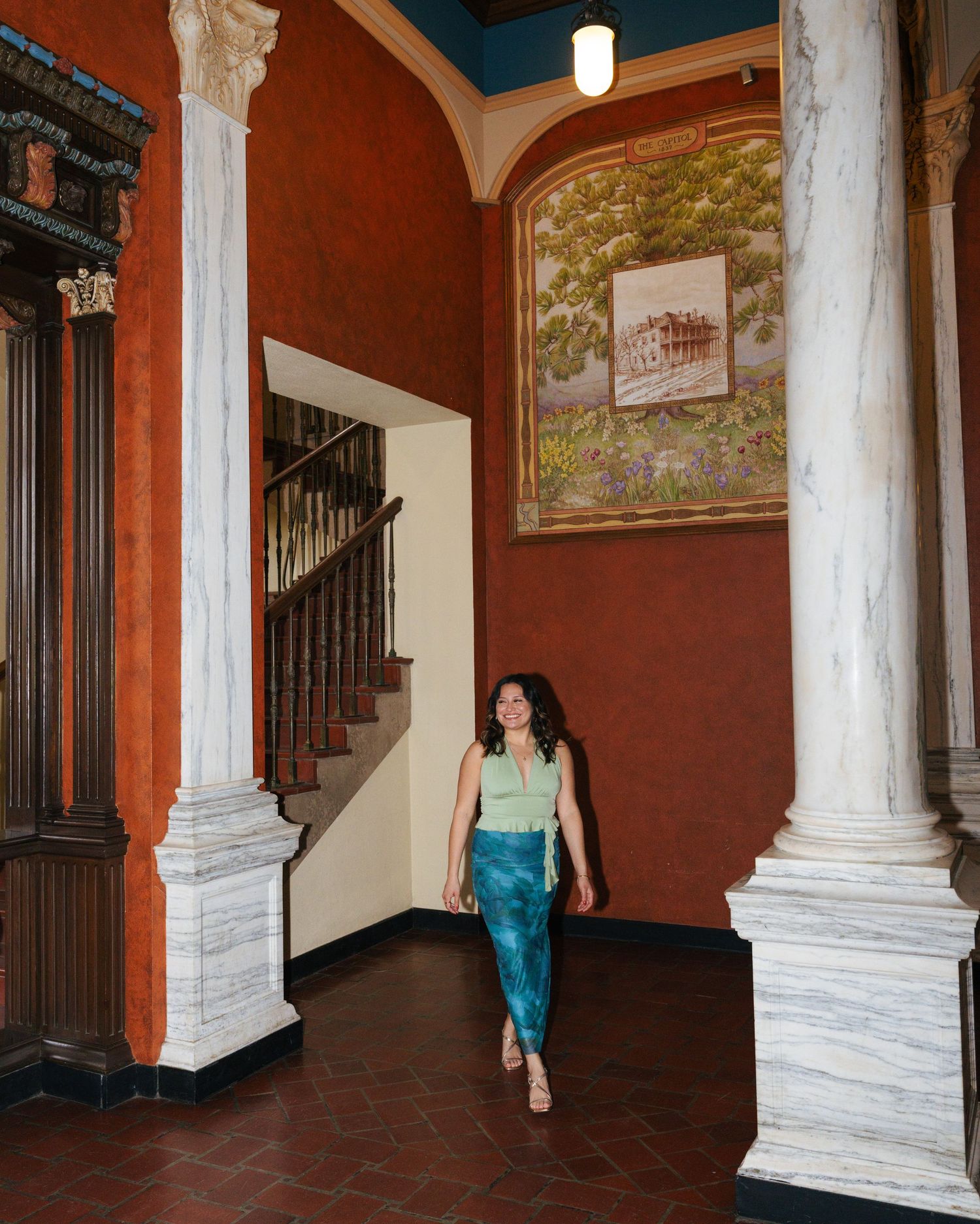 Interior hallway with white marble columns, red walls, and decorative murals features people walking through the space.