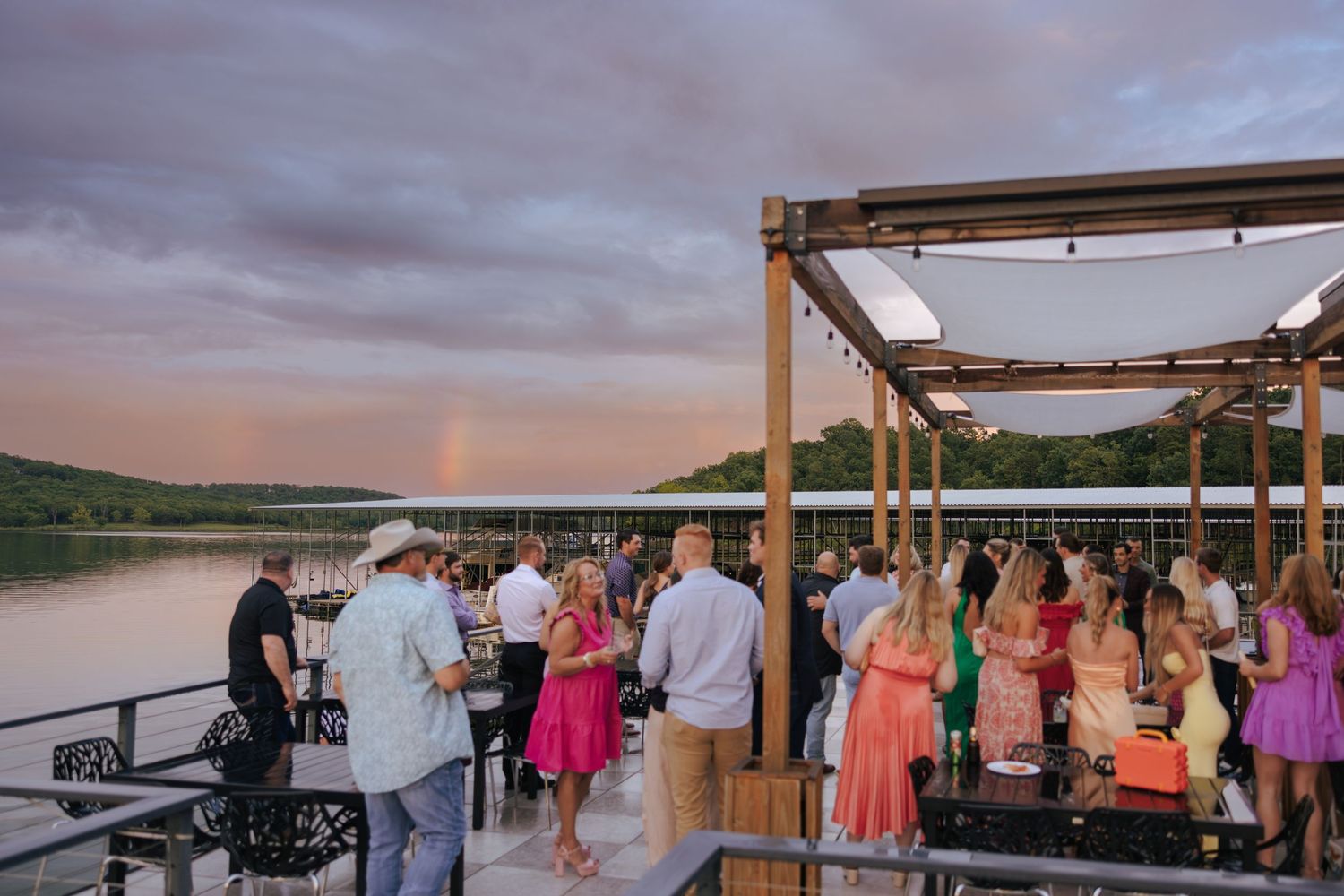 Wedding guests gather on a waterfront deck during sunset as colorful clouds fill the sky above.