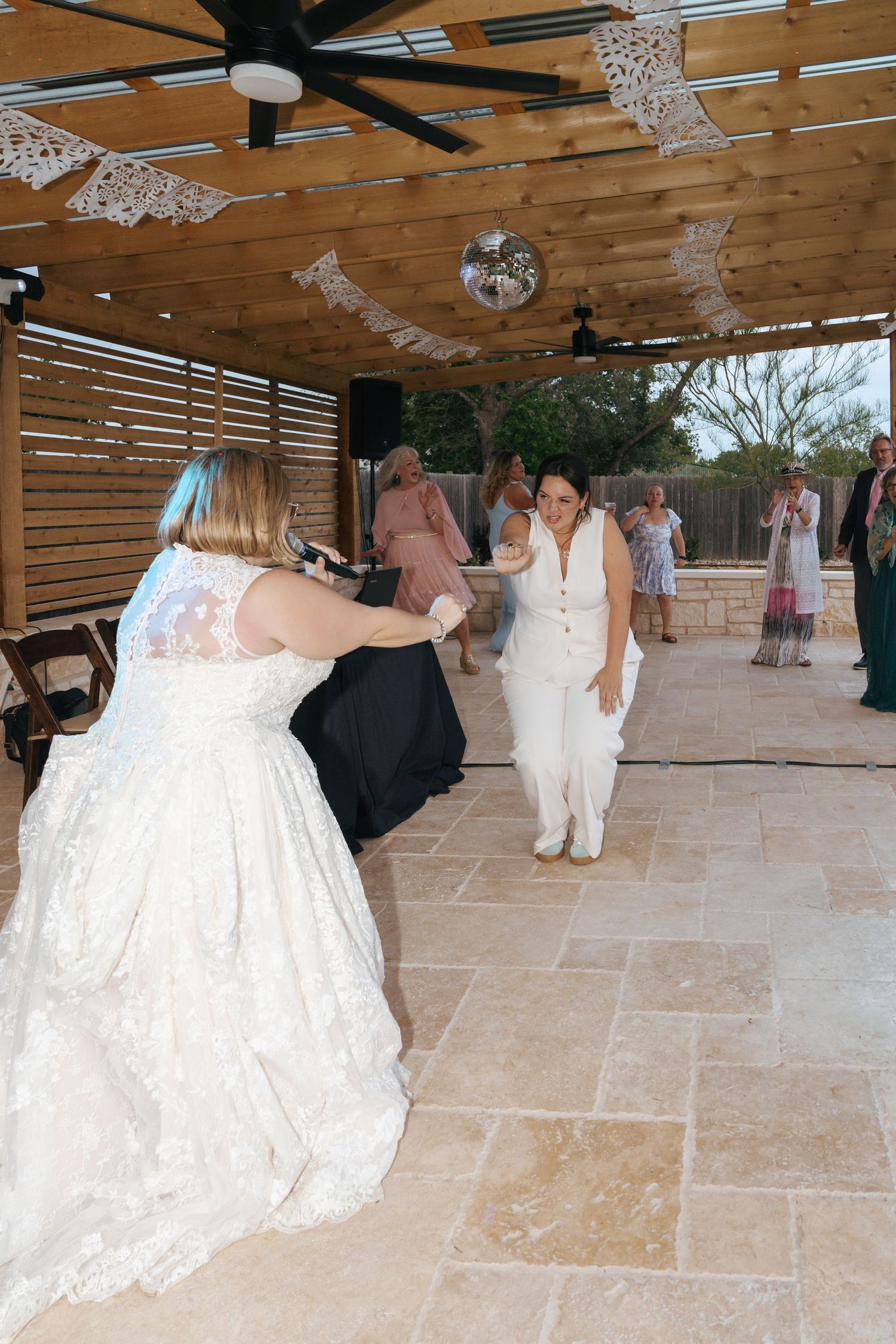 Wedding guests dancing and celebrating under a wooden pergola with decorative lighting and ceiling fans.