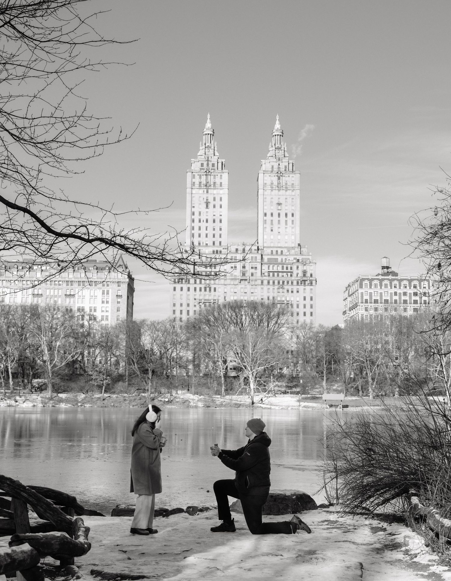 A couple shares a romantic moment by a frozen lake in Central Park with iconic New York City buildings in the background.