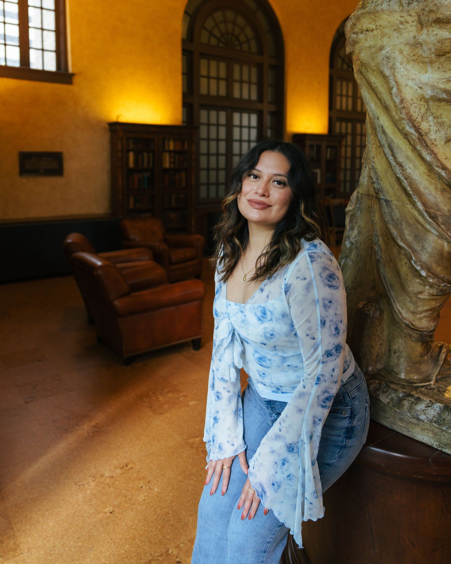 Person in a floral bell-sleeve top and jeans poses in an elegant room with leather chairs and classical decor.