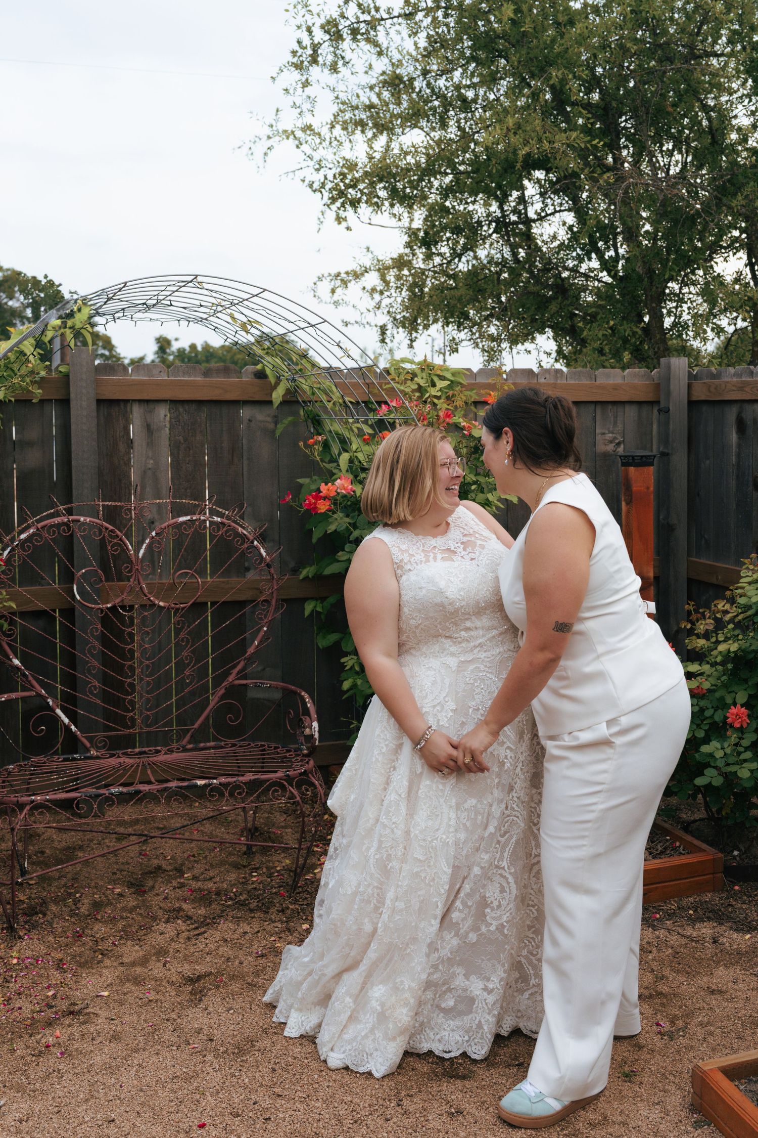 A couple in white dresses share an intimate moment in a garden setting with a wooden fence backdrop.