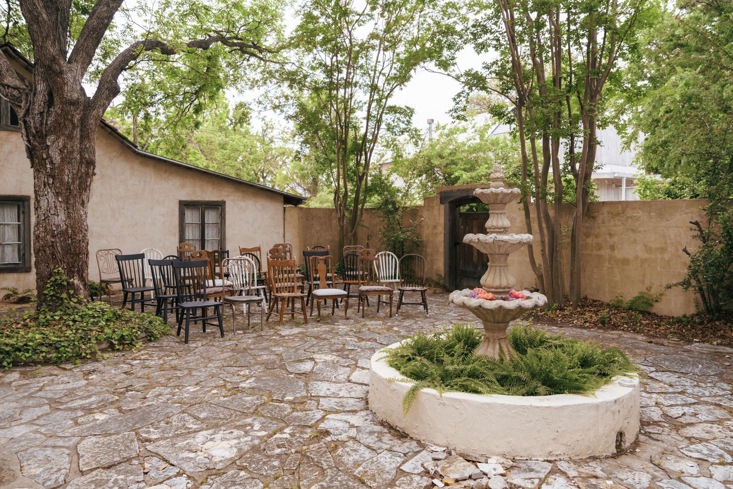 A serene outdoor patio area with stone flooring and rustic wooden chairs arranged under shady trees.