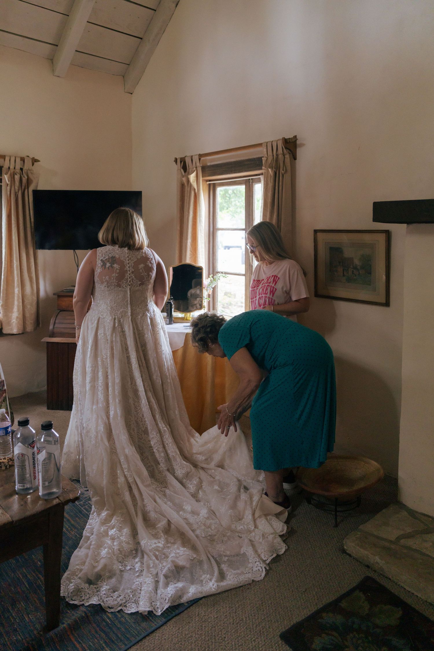 Bridesmaids help adjust the long lace train of a white wedding dress in a sunlit room with beige curtains.
