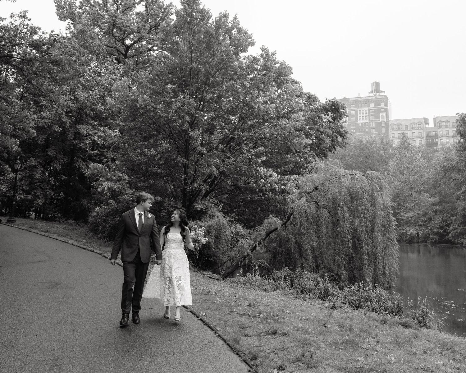 Black and white photo of a couple walking hand in hand along a park path beside a lake, surrounded by lush trees.