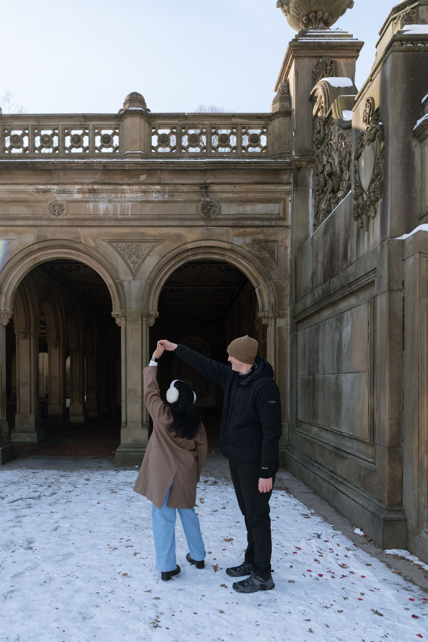 Two people stand in front of a stone archway in winter with snow on the ground at a historic building entrance.