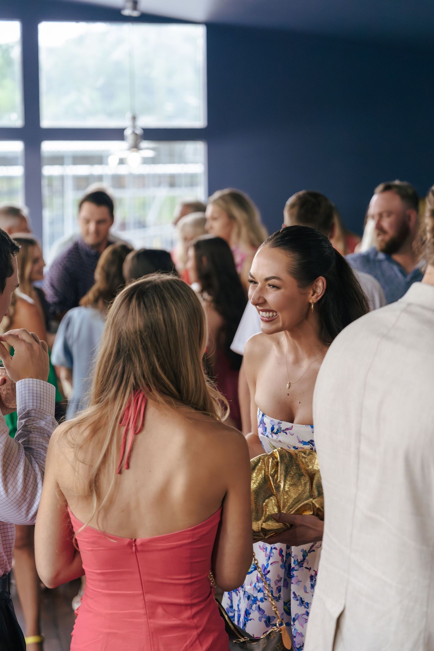 People mingling at an indoor social event with guests wearing summer attire including coral dresses and white shirts.